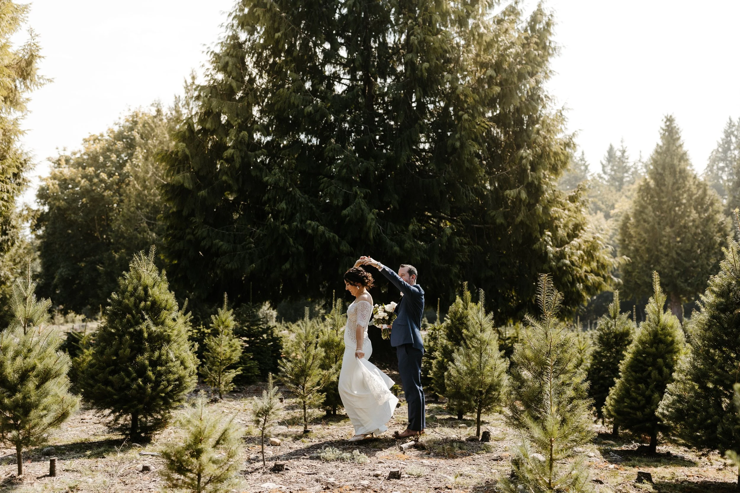 A bride and groom in wedding attire in a forested area, with the groom dancing while holding the bride's hand and she holding a bouquet, surrounded by small pine trees under tall trees with sunlight filtering through.