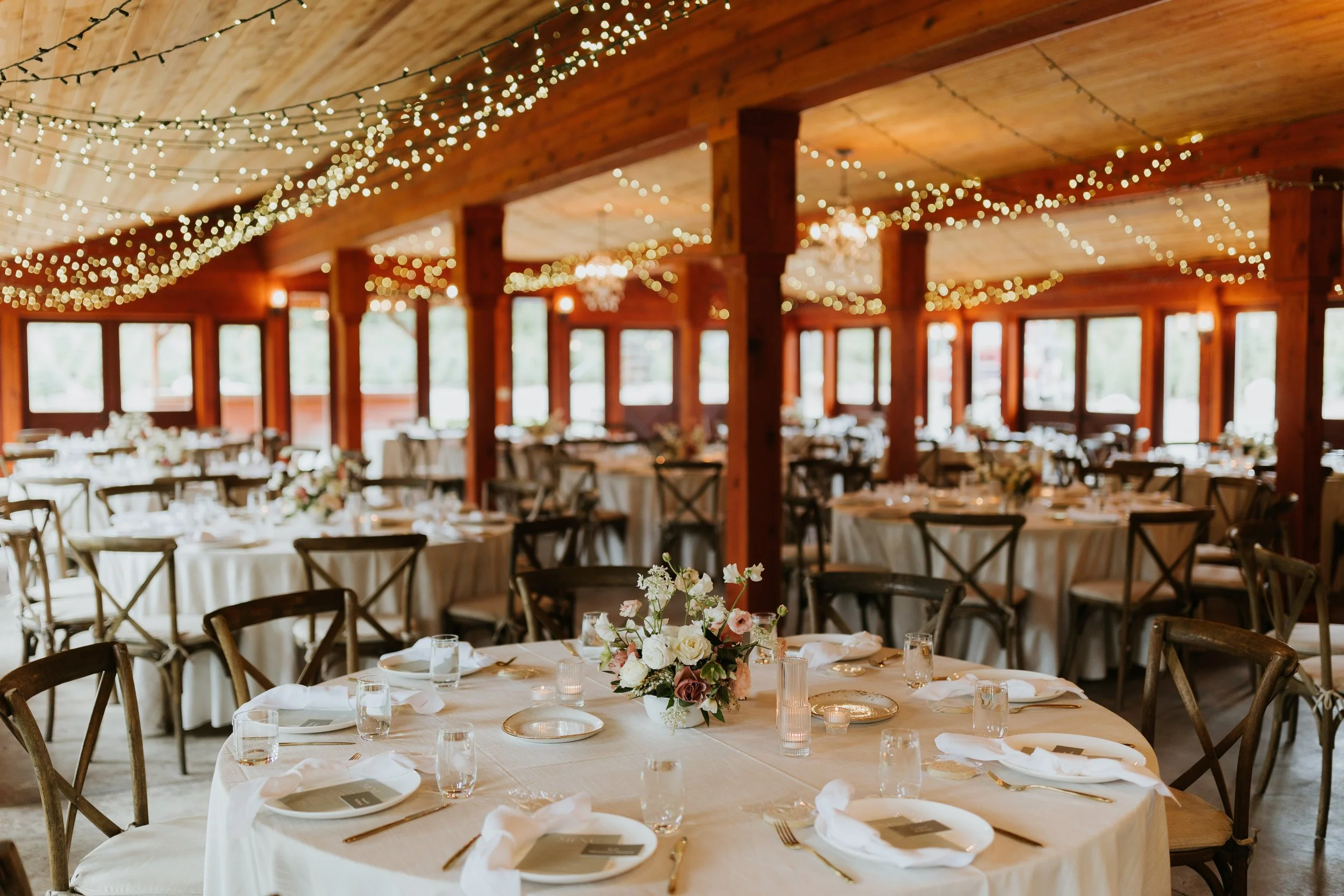 A decorated banquet hall with round tables covered in white tablecloths, arranged with plates, glasses, napkins, and centered with floral arrangements. The hall features wooden walls and ceiling, with numerous string lights hanging, creating a warm,