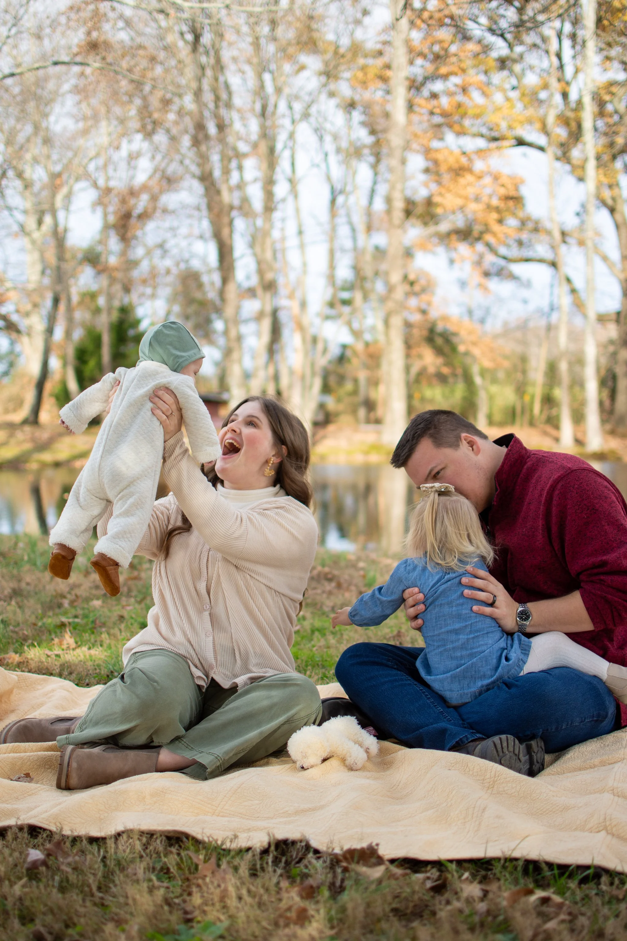 A family of four enjoying a day outdoors near a lake, with two adults and two children, surrounded by trees in autumn, sitting on a blanket on the grass.