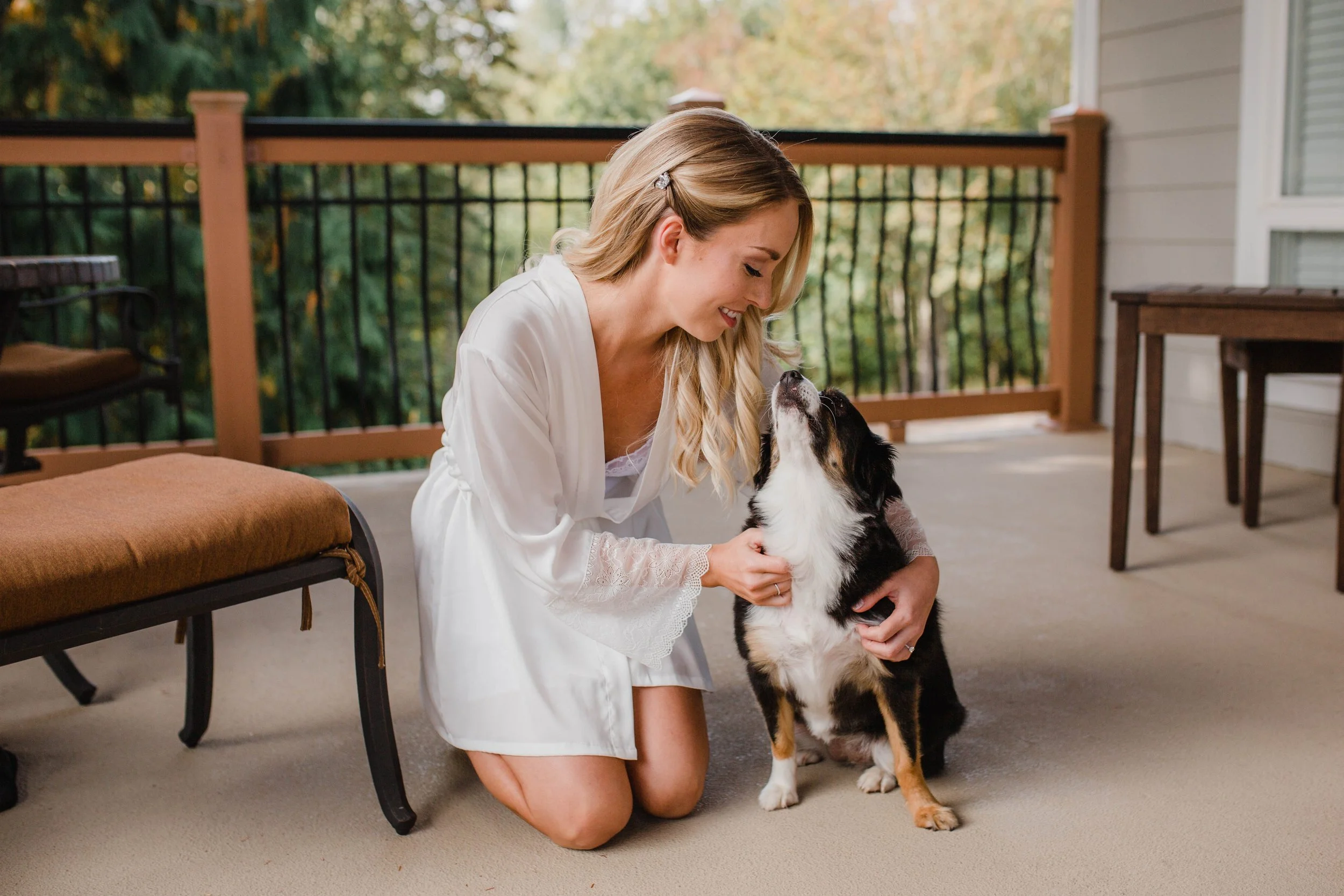 A woman dressed in a white robe smiling while kneeling and petting a black and white dog on a porch with outdoor furniture and a view of trees.