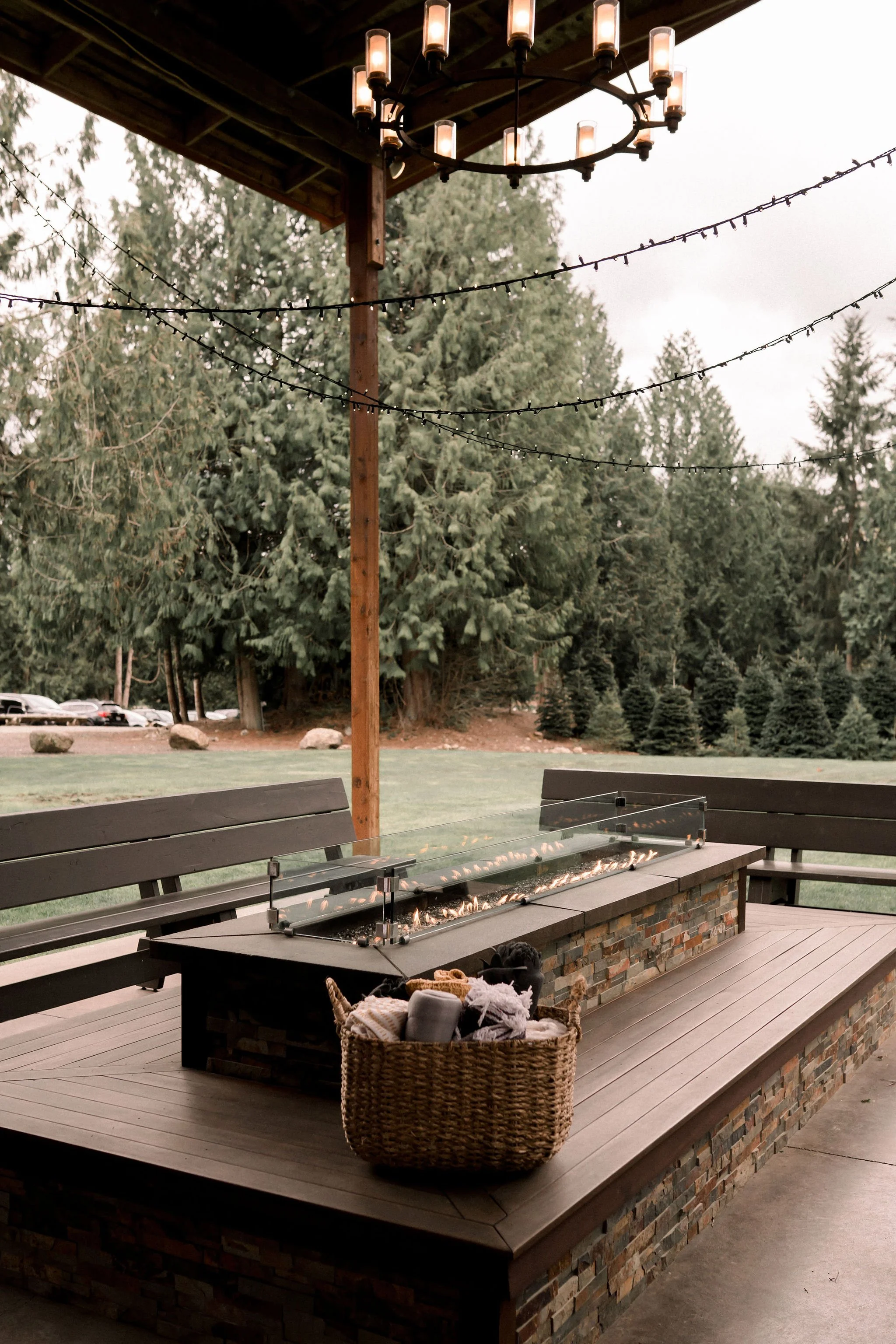 Outdoor patio with a fire pit bar and two benches, surrounded by trees and string lights, under a chandelier.