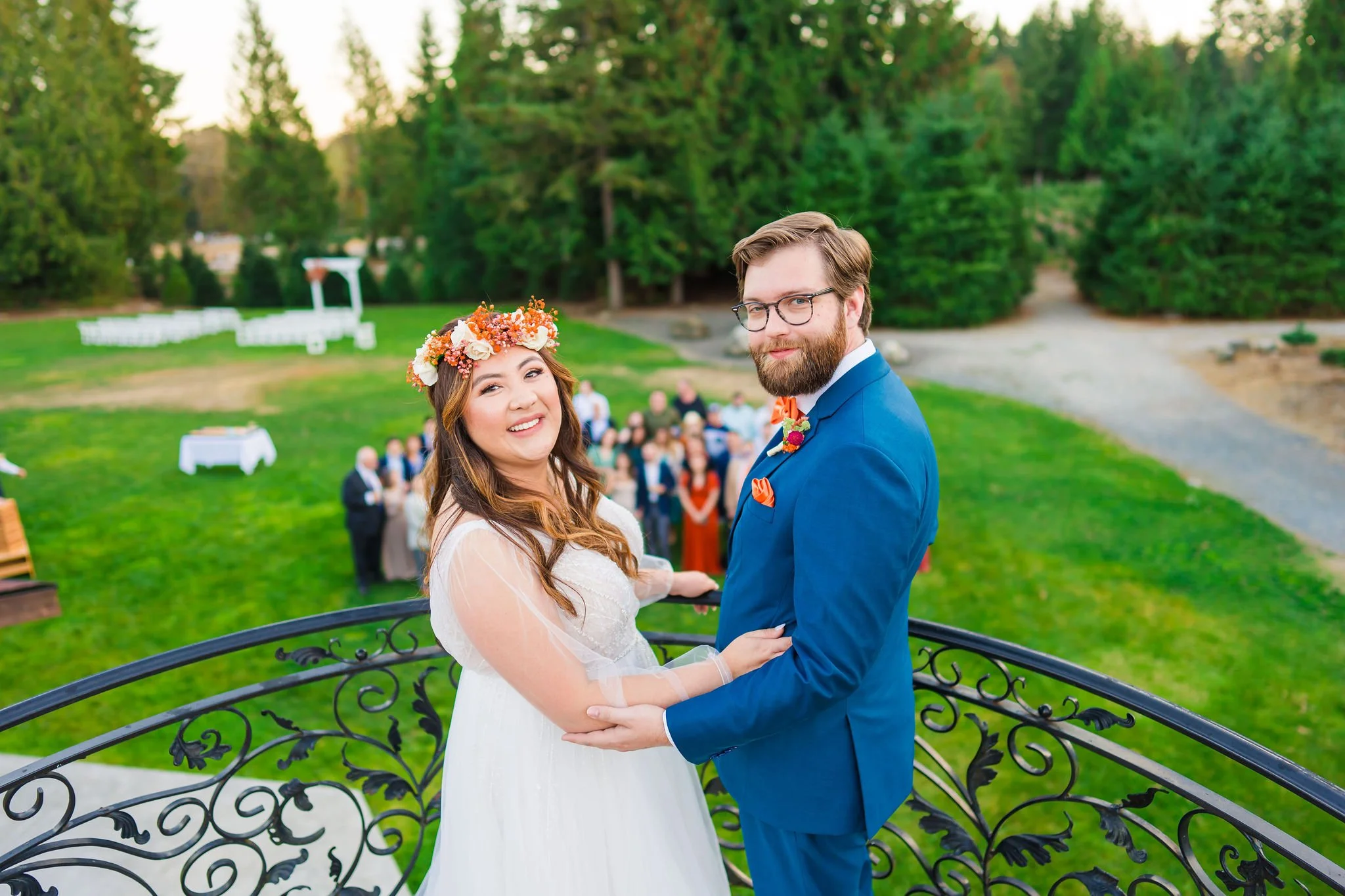A bride and groom holding hands and smiling at a wedding outdoor ceremony with guests in the background, surrounded by lush greenery.