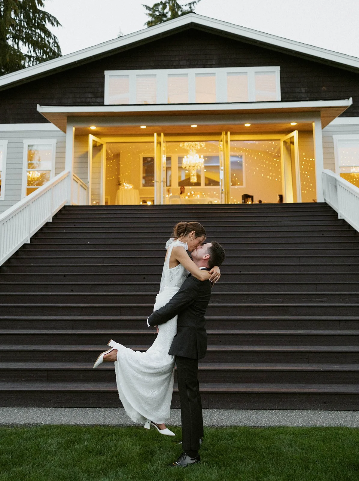 A couple dressed in wedding attire, where the groom is lifting the bride, standing on green grass in front of a large staircase with dark steps and white railings, leading to a house with illuminated windows and glass doors, inside which a chandelier