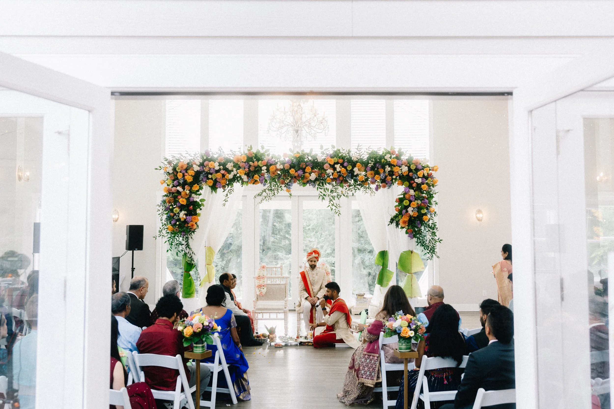 Indoor wedding ceremony with a decorated floral arch, seated guests, and an officiant and groom dressed in traditional attire.