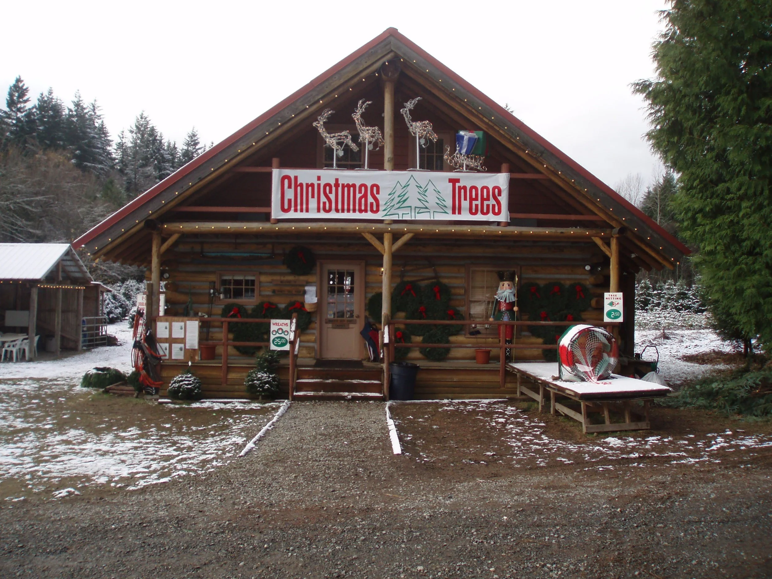 Christmas tree stand with a large sign reading 'Christmas Trees', decorated with wreaths, Christmas lights, and festive decorations, set against a snowy outdoor setting.