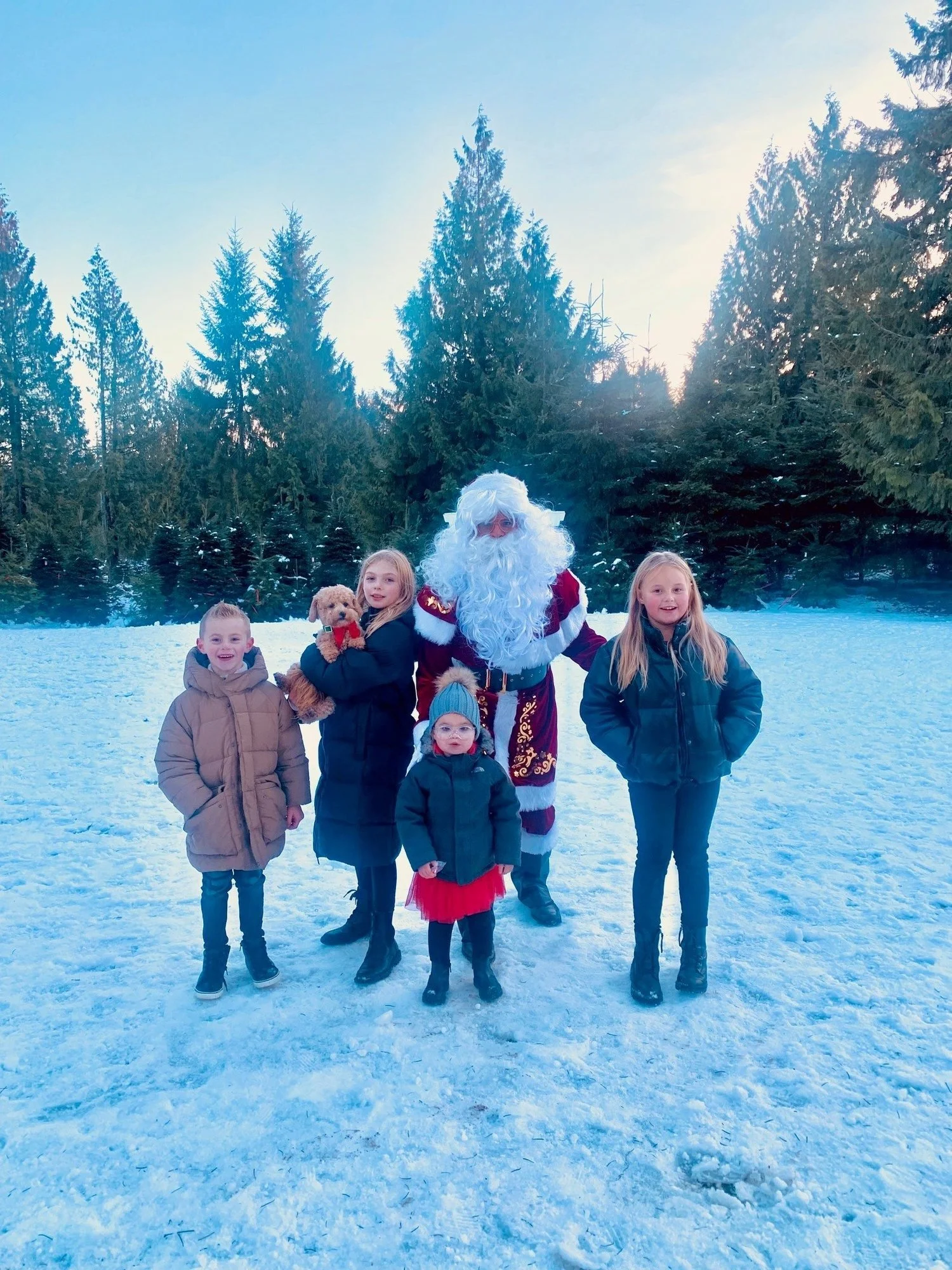 Children posing with a person dressed as Santa Claus in a snowy outdoor setting with evergreen trees in the background.