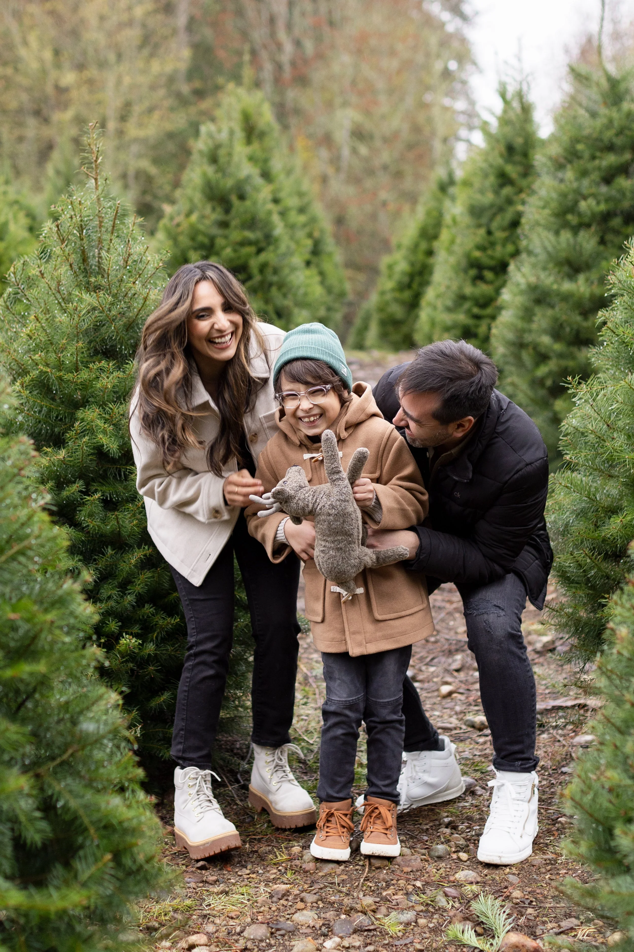 A family of three with a woman, a man, and a young boy are smiling and playing with a stuffed animal in a Christmas tree farm surrounded by evergreen trees.