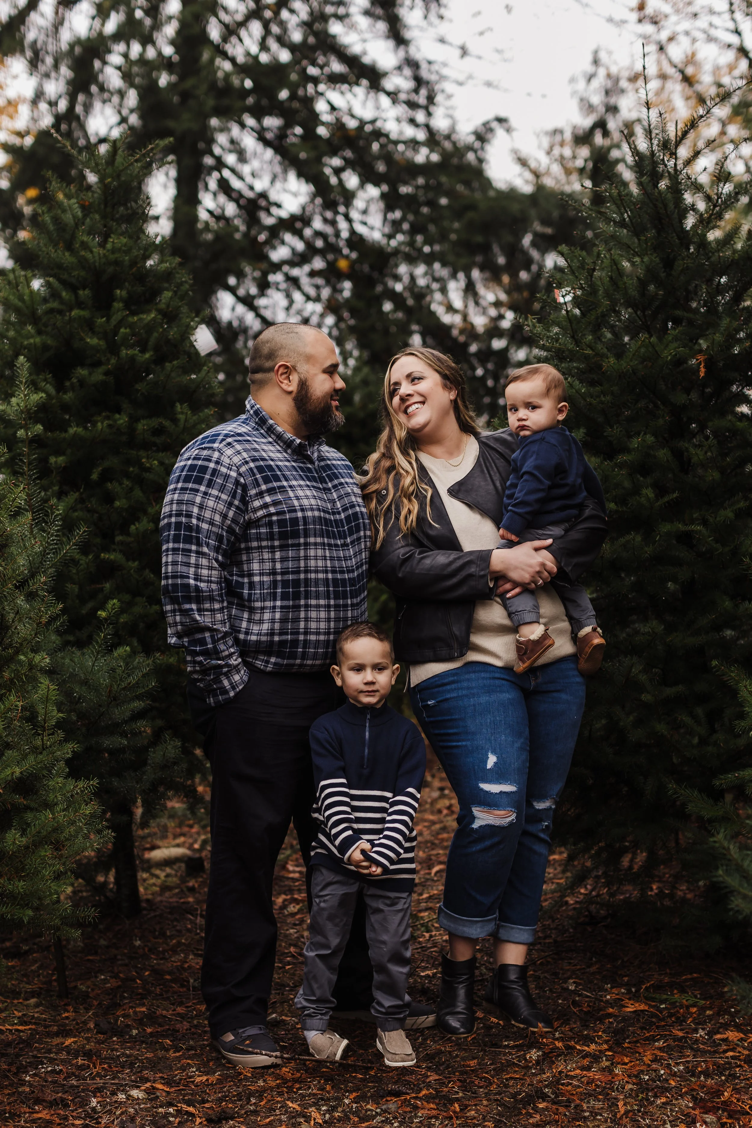 A family of four stands among Christmas trees in an outdoor setting, smiling and looking at each other.