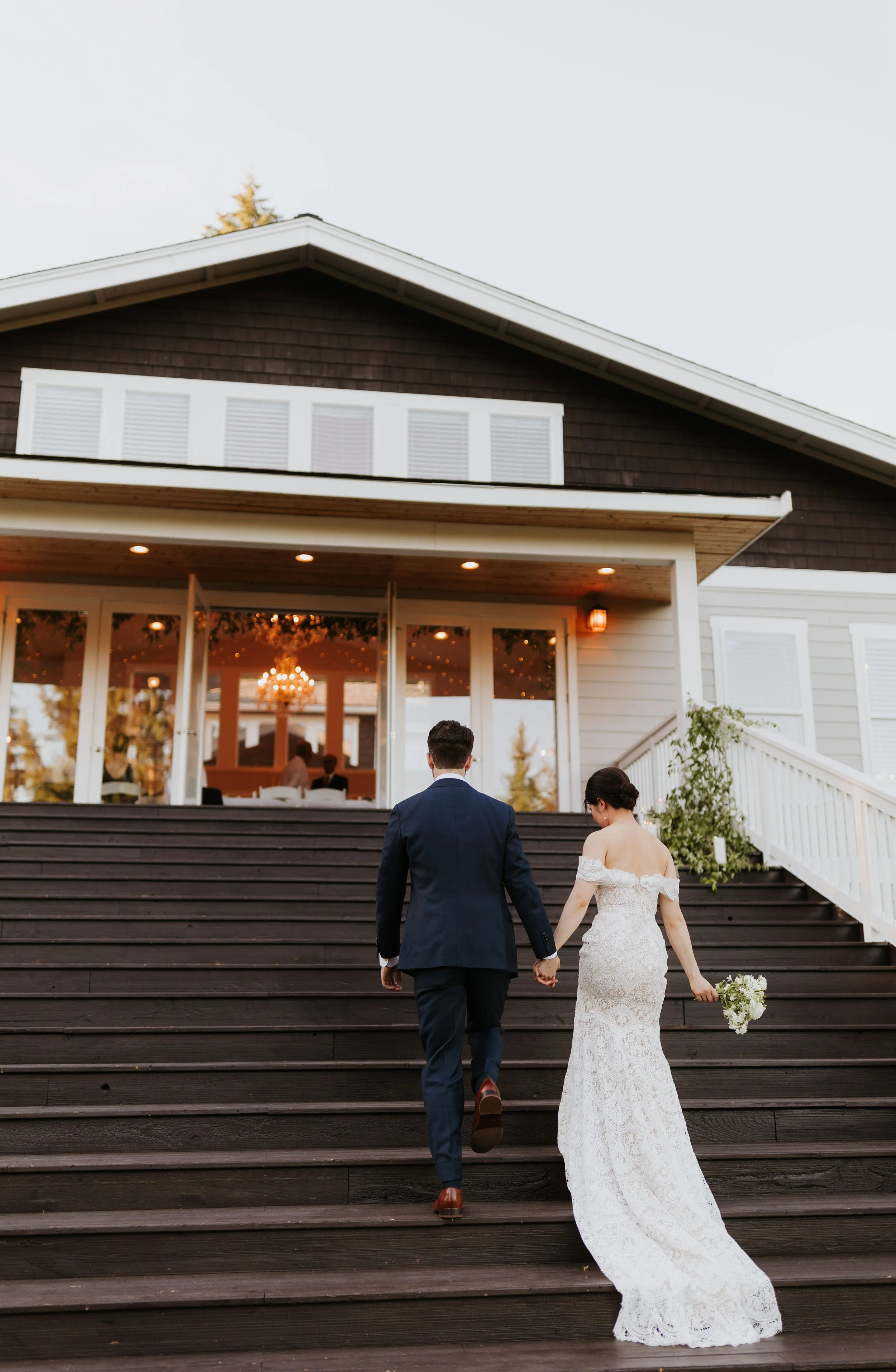 A bride and groom ascending outdoor wooden stairs toward a house, holding hands. The bride is in a white lace wedding gown holding a bouquet, and the groom is in a dark suit. The house has large windows and a welcoming porch with warm lighting.
