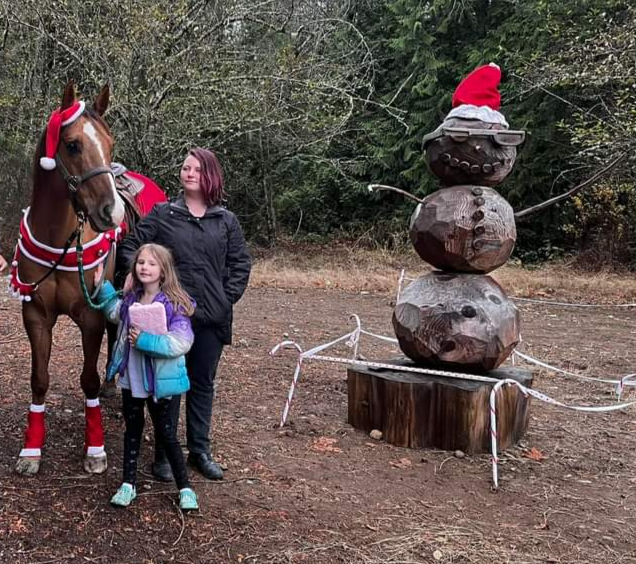 Two children and an adult standing next to a decorated horse in an outdoor wooded area. The horse has a red Santa hat, a red and white stripe vest, and red leg wraps. There is a large wooden snowman sculpture with a Santa hat and scarf nearby.