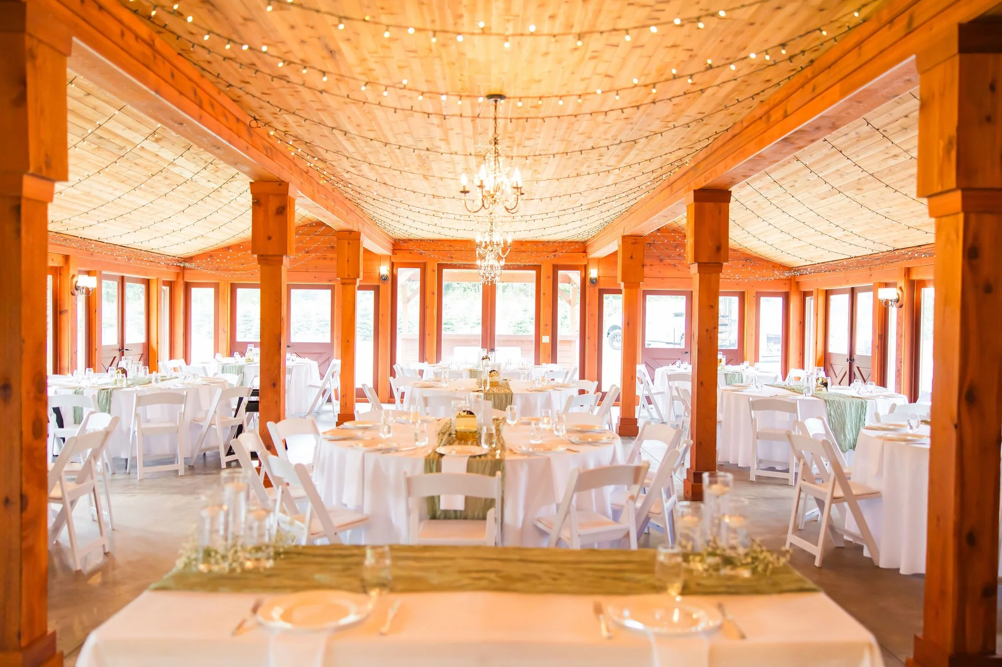 Wedding reception hall decorated with string lights, chandeliers, and round tables with white tablecloths and chairs.