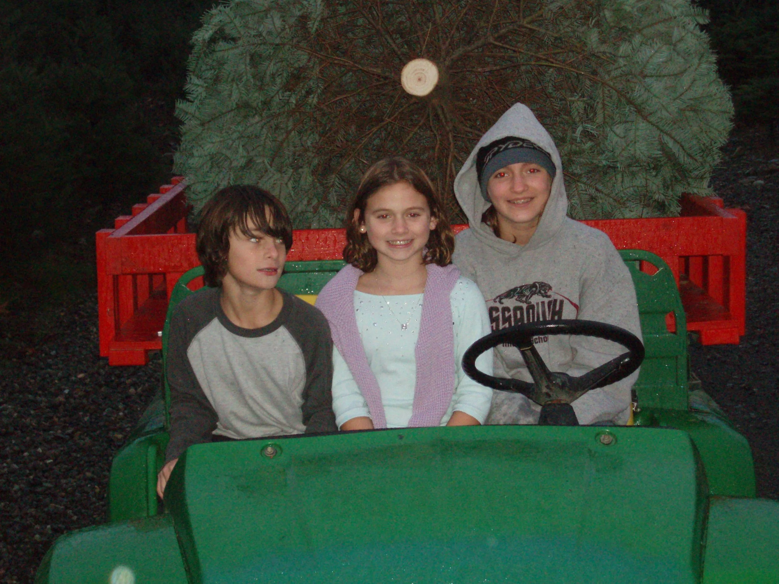 Three children sitting in a green toy vehicle with a large Christmas tree in the background.