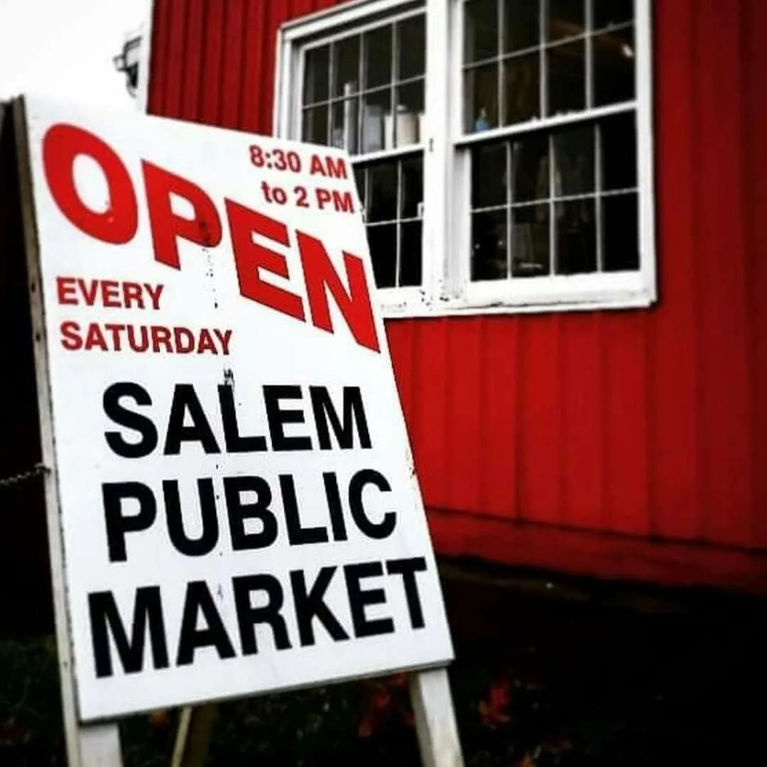 A sidewalk sign outside a red building with white-framed windows, indicating the Salem Public Market is open every Saturday from 8:30 AM to 2 PM.