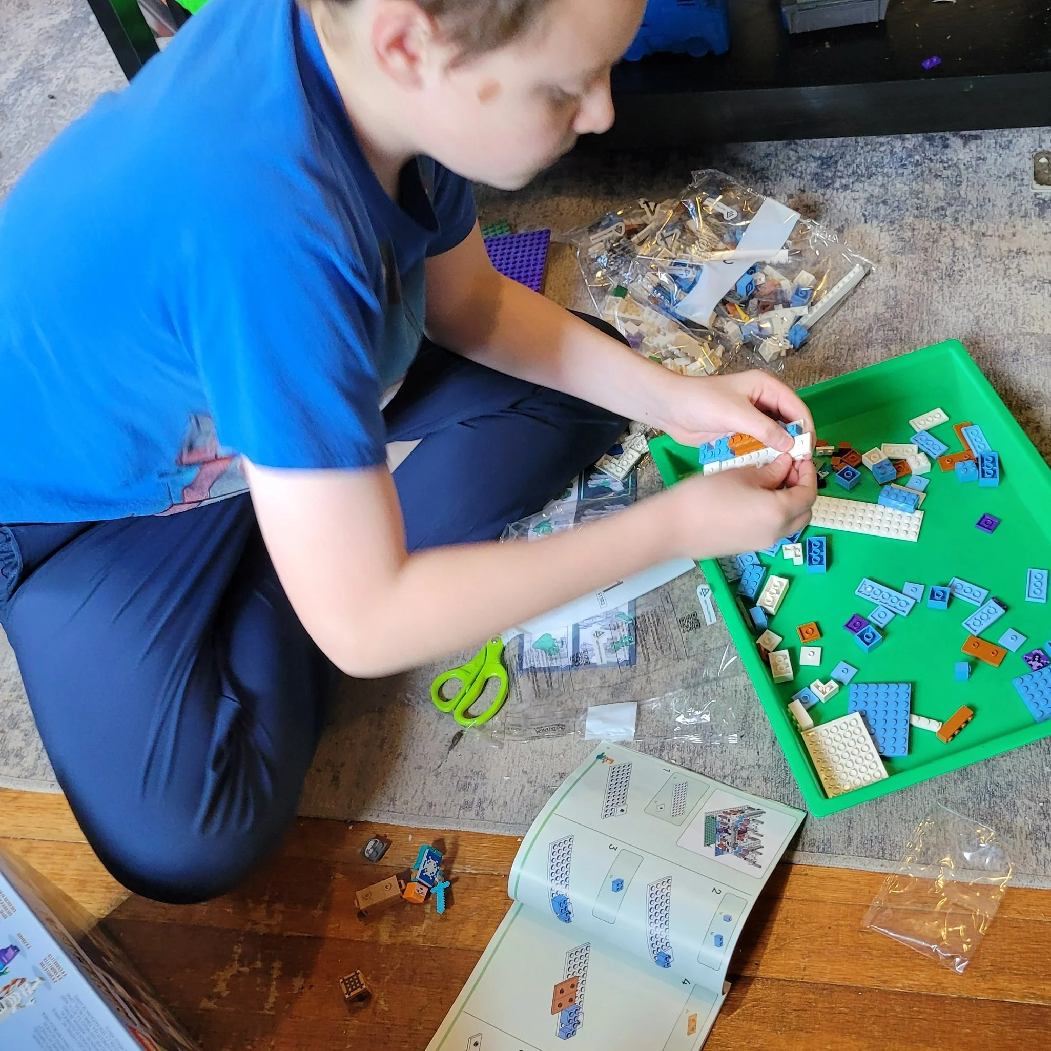 A young boy is sitting on the floor assembling a Lego set. He is wearing a blue T-shirt and dark pants. There are various Lego pieces in a green tray, scattered around him, and an instruction booklet open in front of him. Scissors and other small Lego pieces are also nearby.