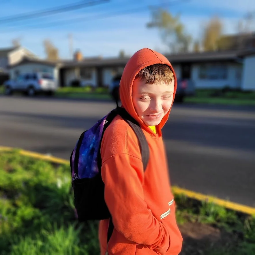 A young boy with a backpack wearing an orange hoodie with the hood up, standing outdoors on a sidewalk with a residential street in the background, smiling with eyes closed.