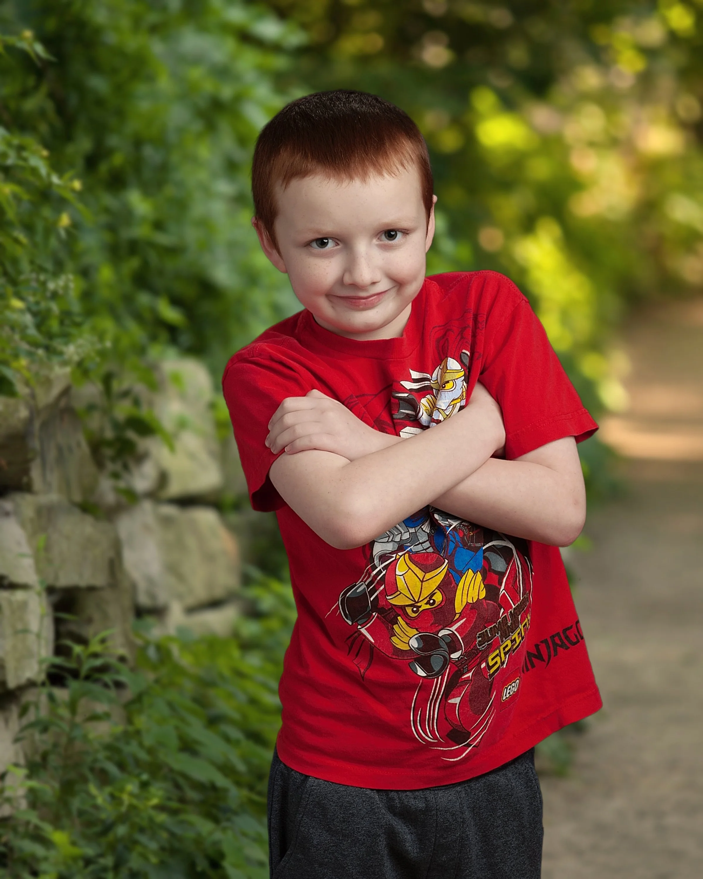 A young boy with short brown hair and blue eyes standing outdoors on a pathway, crossing his arms and smiling, wearing a red Lego Ninjago T-shirt.