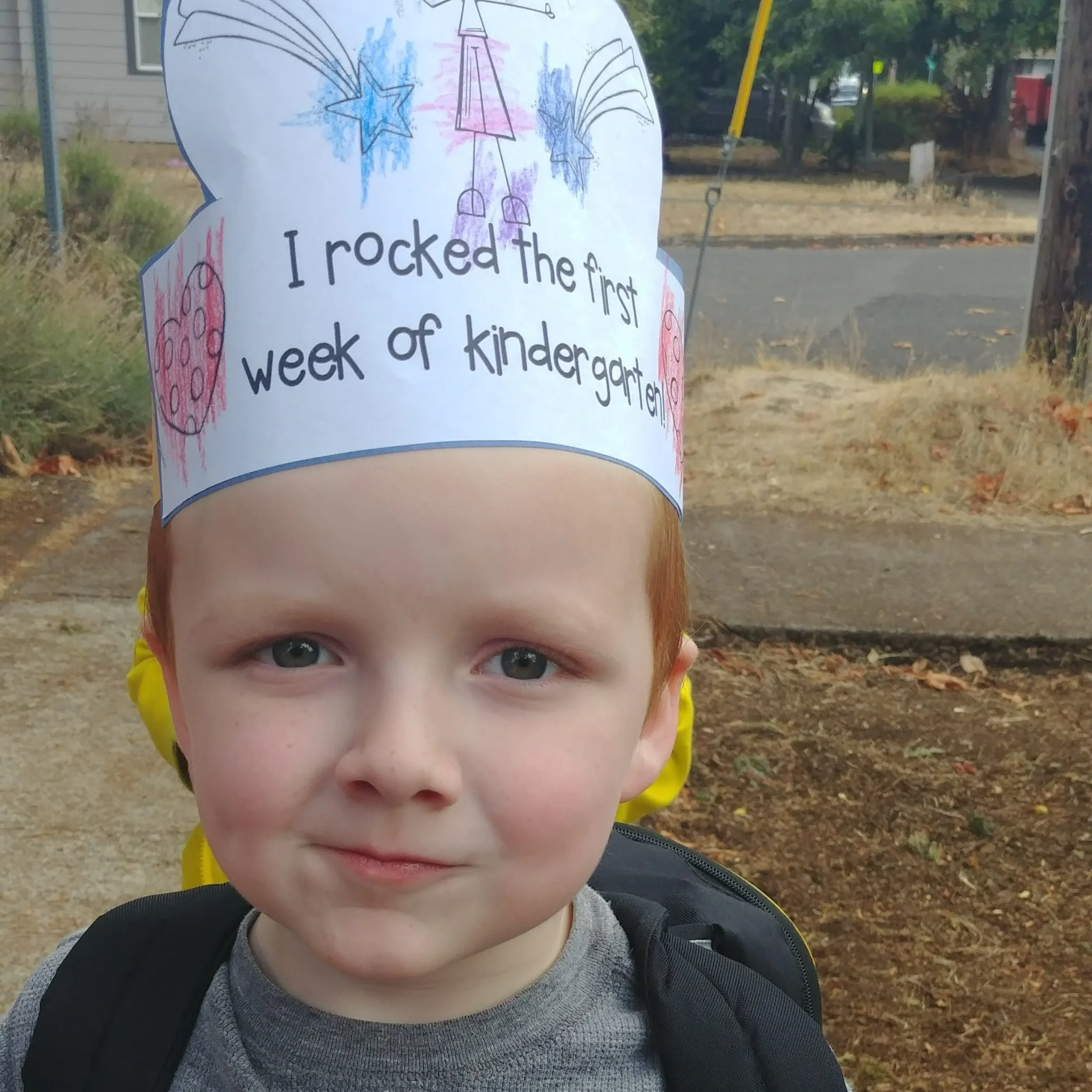 Young boy with a paper crown on his head that says 'I rocked the first week of kindergarten,' standing outdoors near a sidewalk and trees.