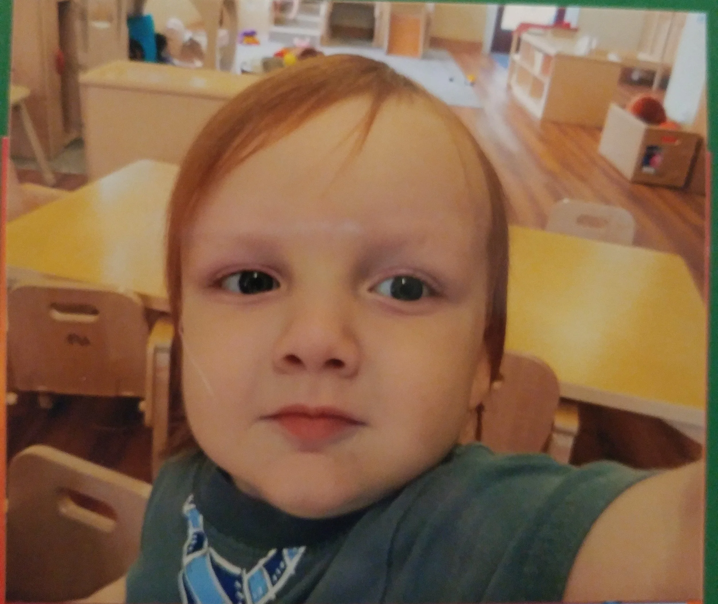 Close-up of a young boy with reddish hair and big eyes taking a selfie in a classroom with yellow tables and wooden chairs.