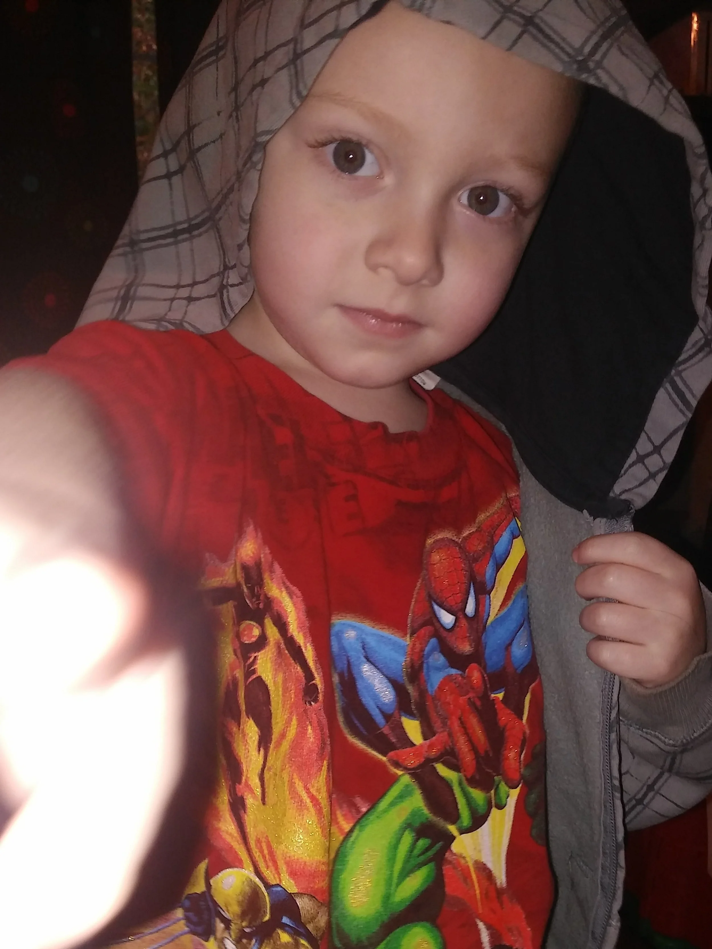 A young boy with big eyes and a slight smile, wearing a red Spider-Man t-shirt, lying on a pillow with a gray plaid pattern, with a thumb partially covering the camera in a close-up selfie.