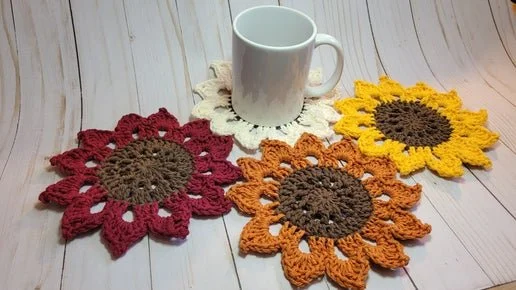 Four crocheted sunflower coasters in red, yellow, orange, and white with a plain white mug on a light wooden surface.
