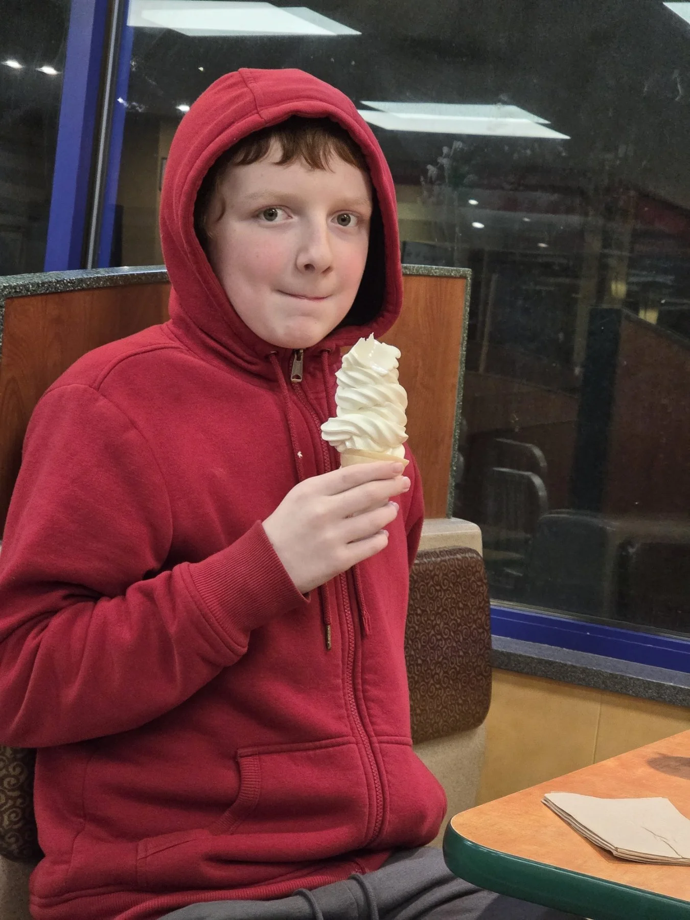 A boy in a red hoodie holding a large vanilla soft serve ice cream cone inside a restaurant.
