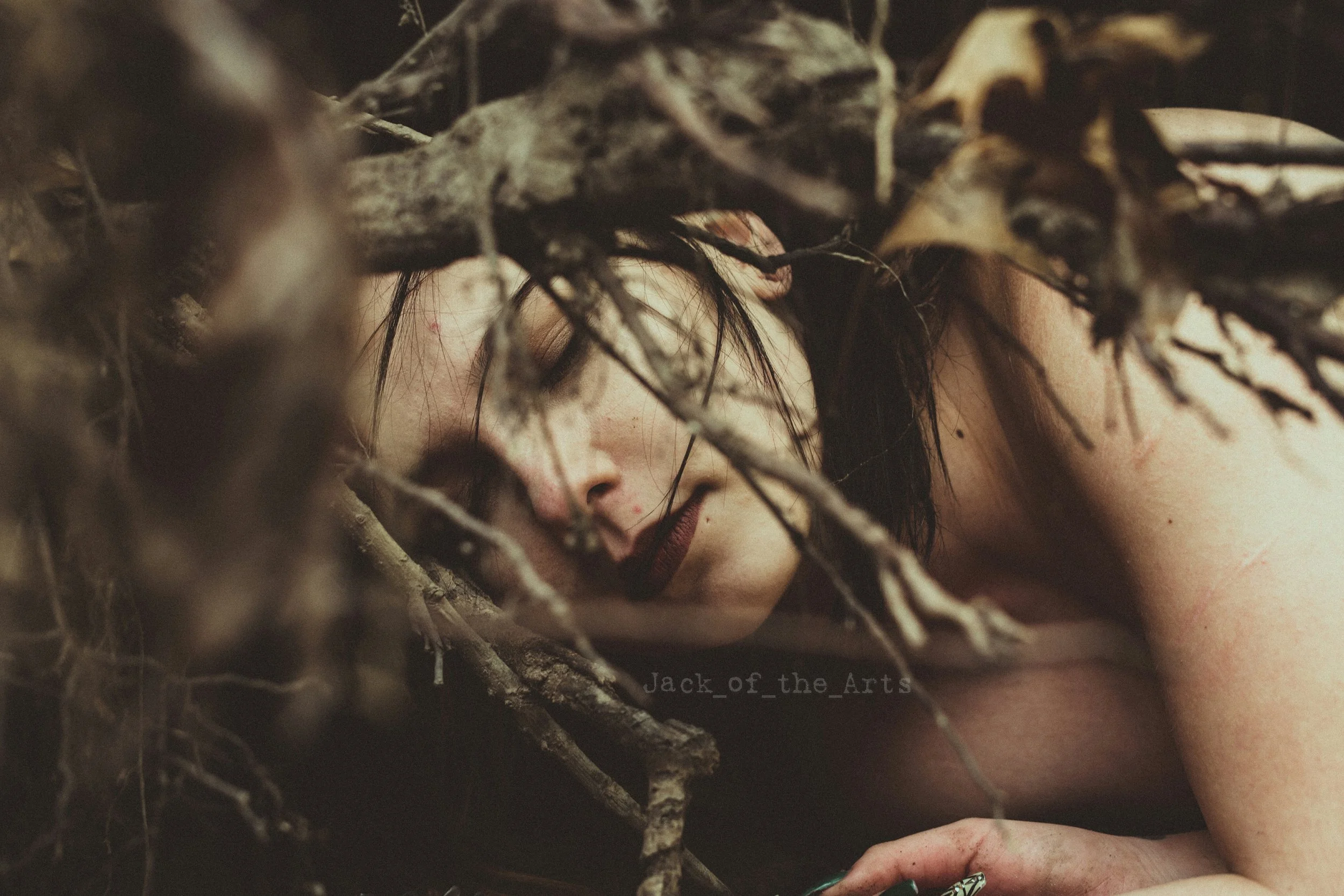 A woman with dark hair and makeup, lying among tree branches and leaves, with her eyes closed and a serene expression.
