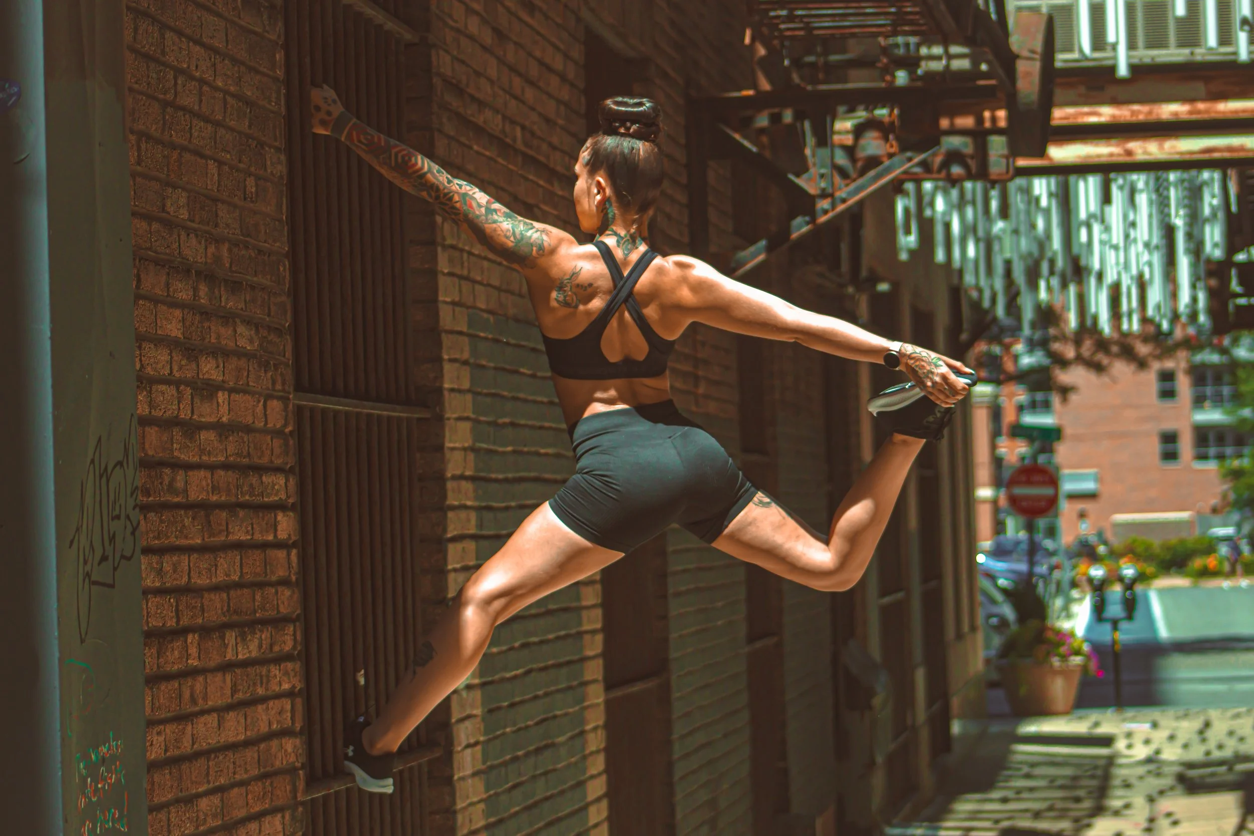 A woman with tattoos in athletic clothing performing a wall jump on a brick building sidewalk.