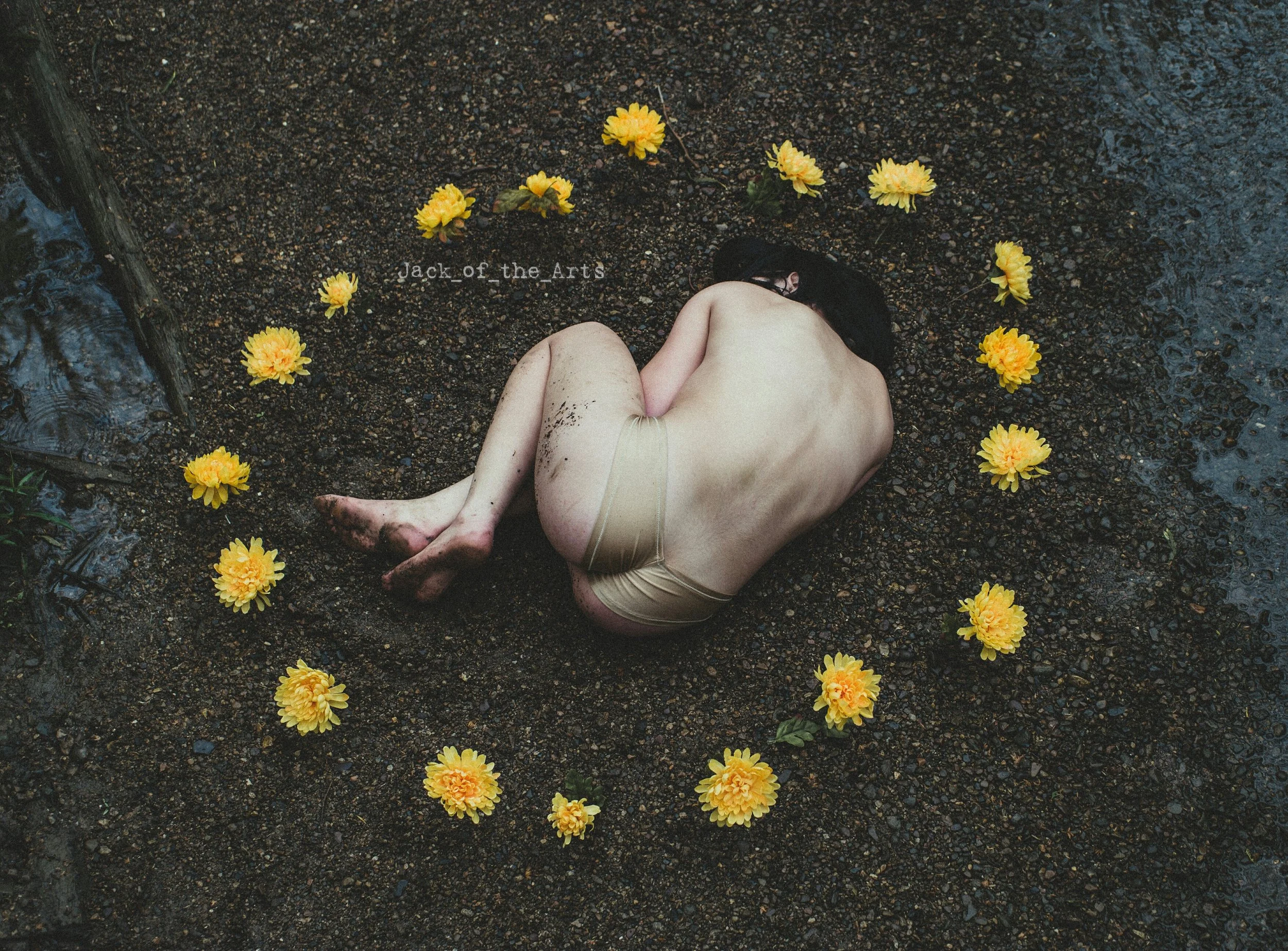A naked person with dark hair lying on wet, dark soil, surrounded by yellow and orange flowers, with mud on their legs.
