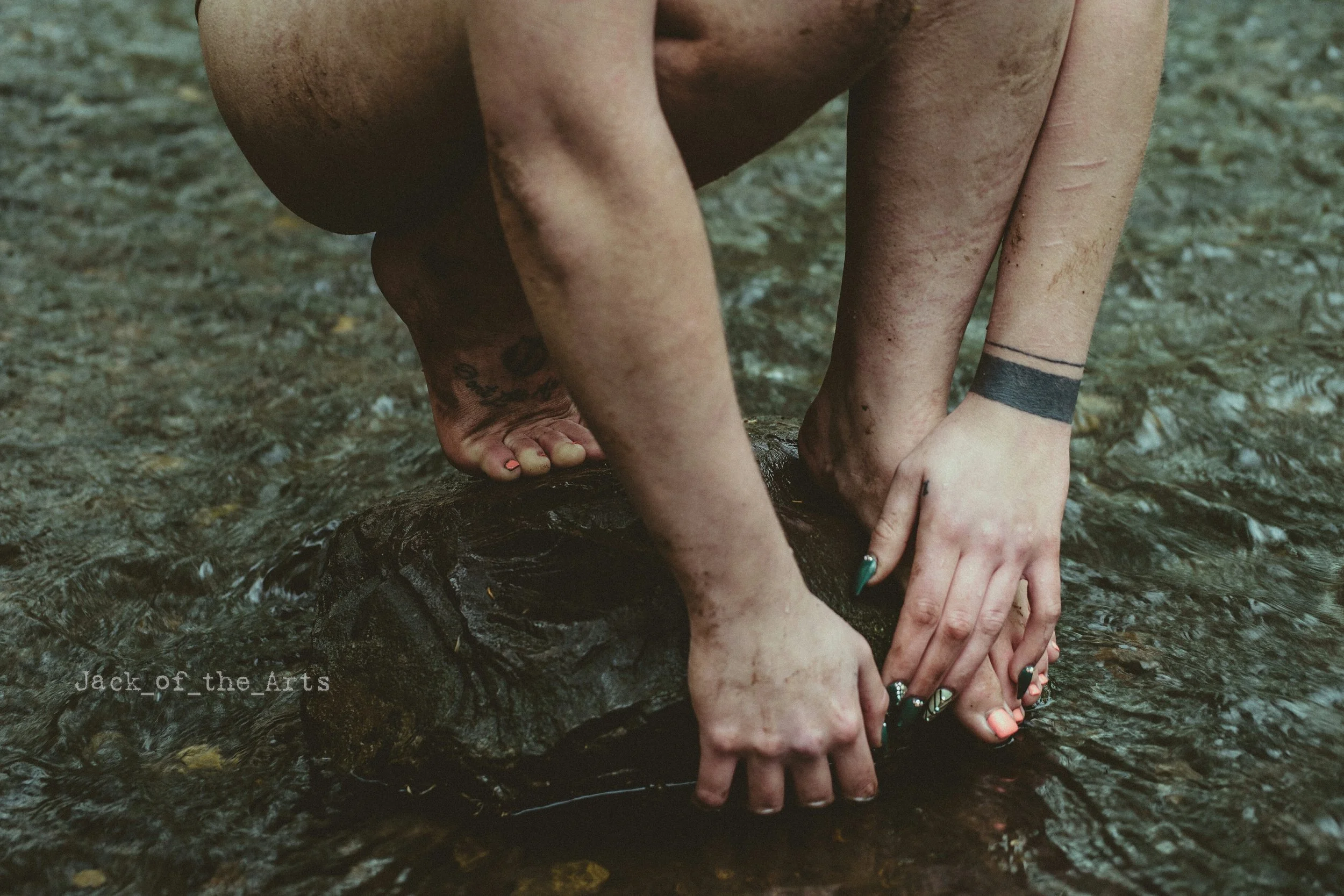 Person crouching in a shallow stream, touching a rock with hands and feet, with visible tattoos and brightly colored nails.