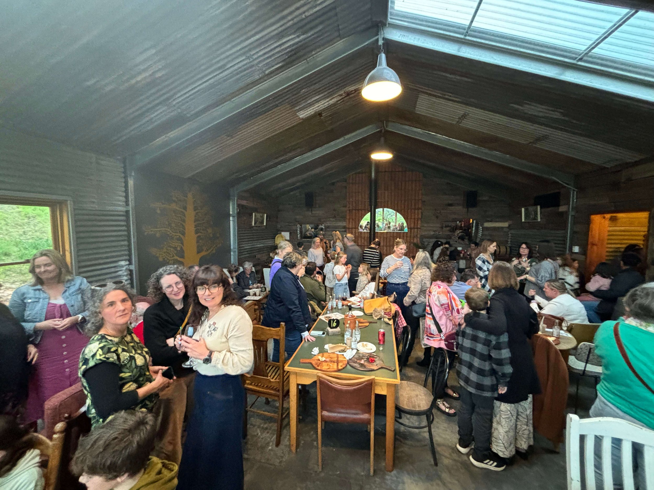 A large group of people gathered inside a rustic barn with metal walls, wooden accents, and hanging lights, socializing and enjoying food and drinks.
