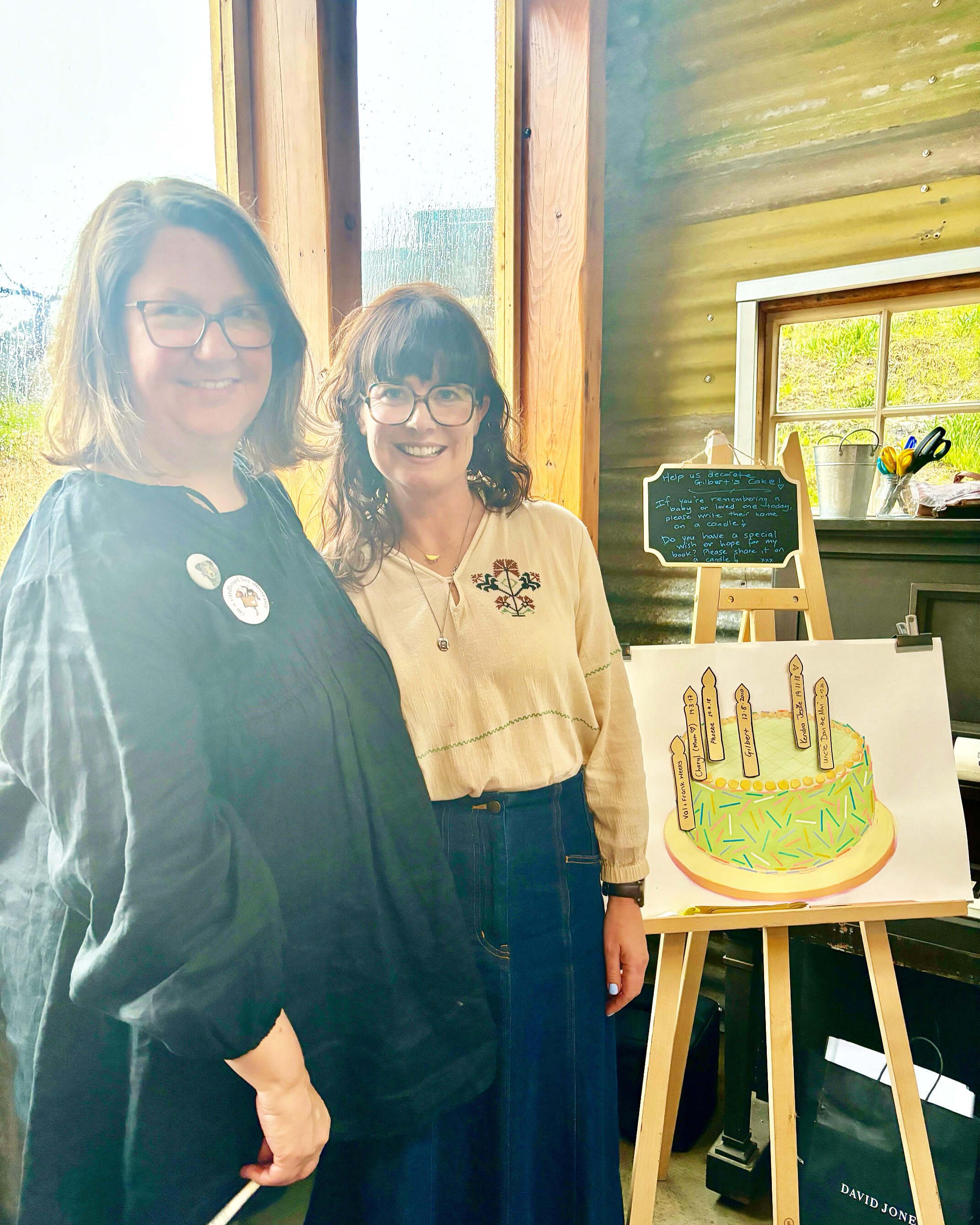 Two women standing indoors near a colorful birthday cake illustration on a sign and a small chalkboard with handwritten message, both smiling at the camera.