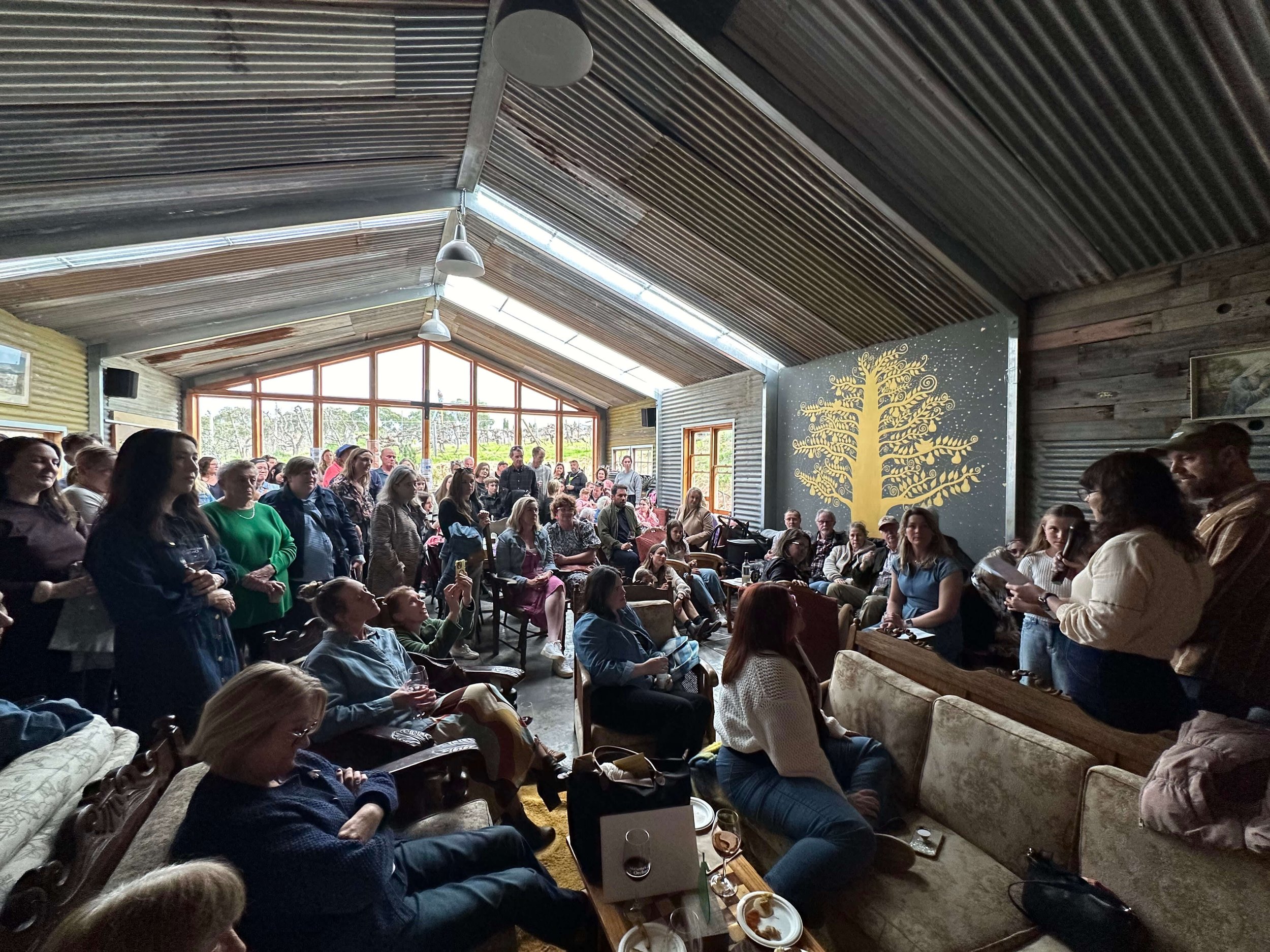 Crowd gathered in a rustic, barn-like interior with a large window and a decorative golden tree mural, attending an event or presentation.
