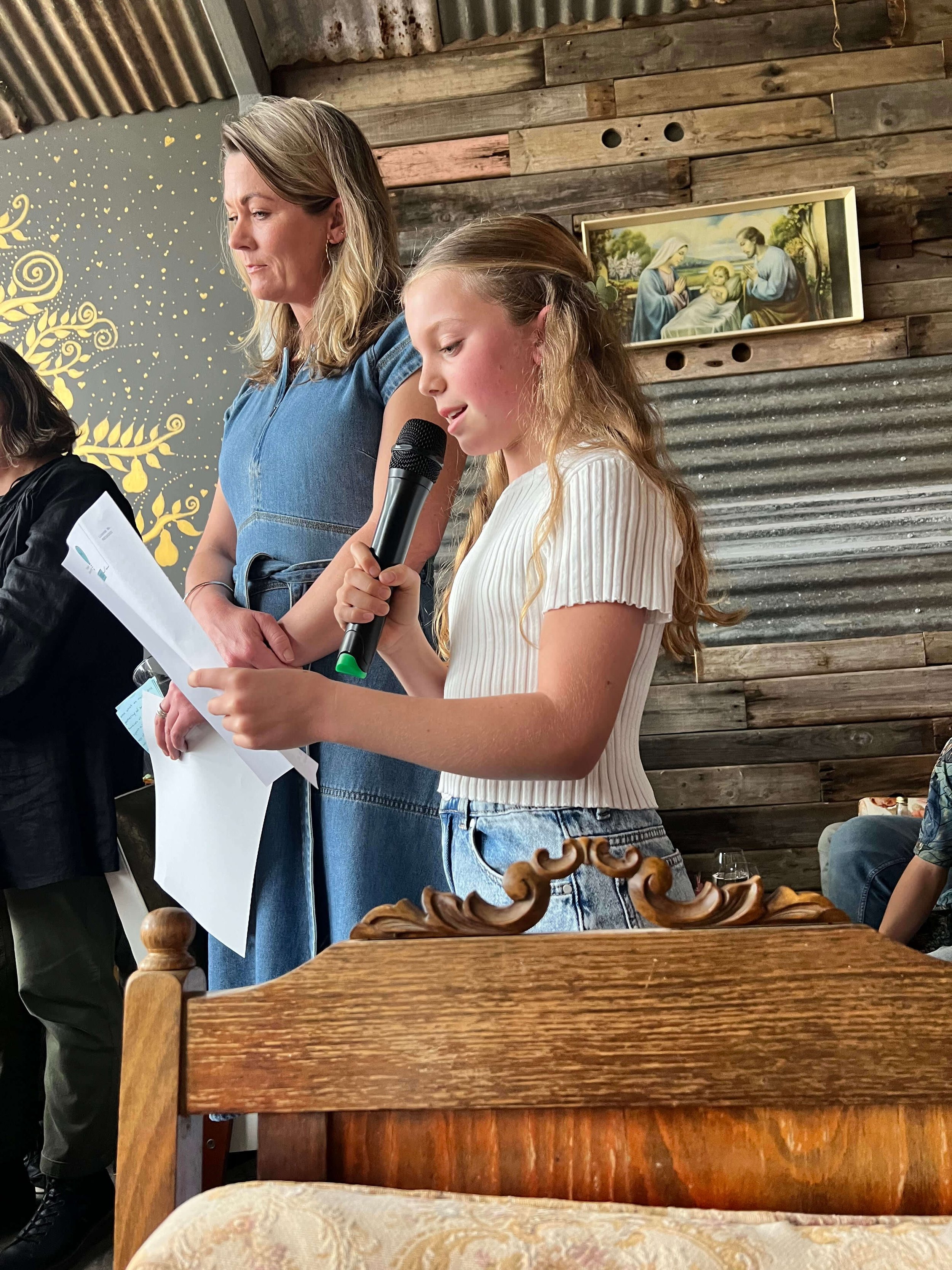 A young girl with long, wavy blonde hair holding a microphone and reading from a sheet of paper, standing next to an adult woman with shoulder-length blonde hair in a denim dress, in a rustic interior with wooden walls and religious art.