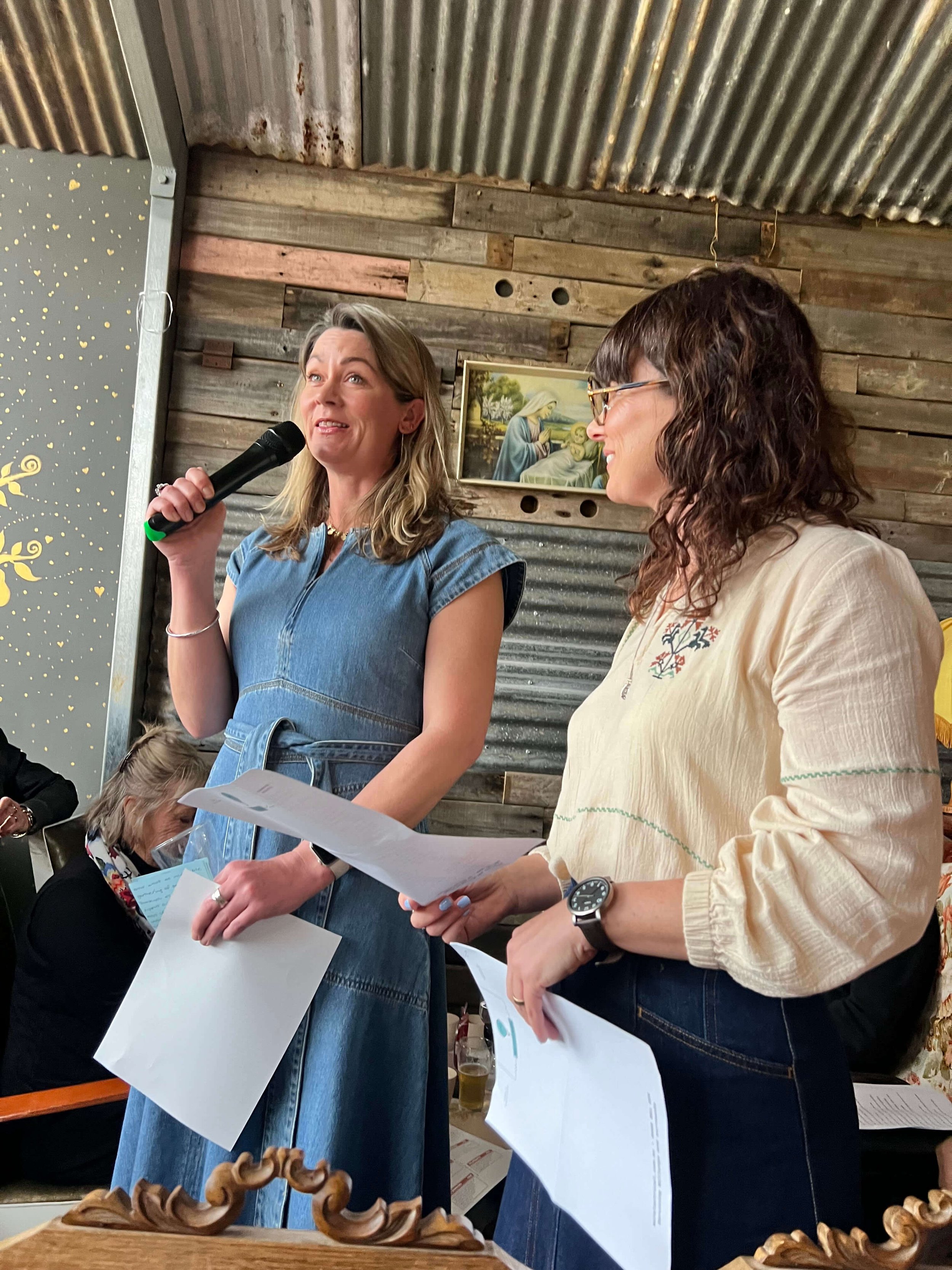 Two women standing and speaking at an indoor event, with one holding a microphone and papers, in front of a rustic wooden wall with religious artwork.
