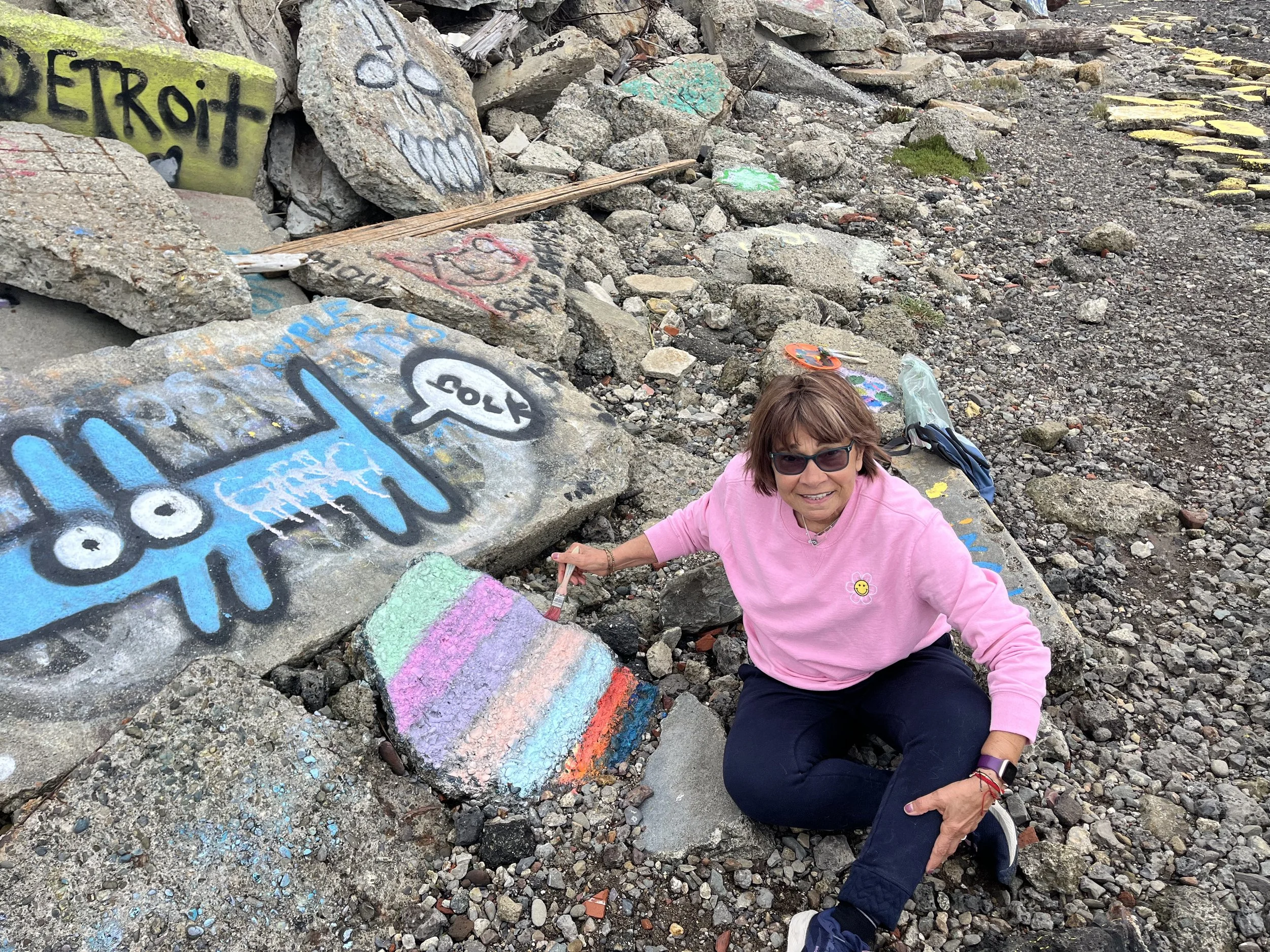 A woman in a pink sweatshirt and sunglasses is sitting on a rocky beach, painting a large inukshuk figure on the rocks with bright colors. There are graffiti and colorful designs on the rocks around her.