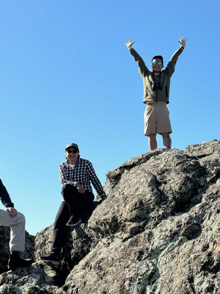 Two people on rocky terrain, one standing with arms raised and the other sitting on a rock, under a clear blue sky.