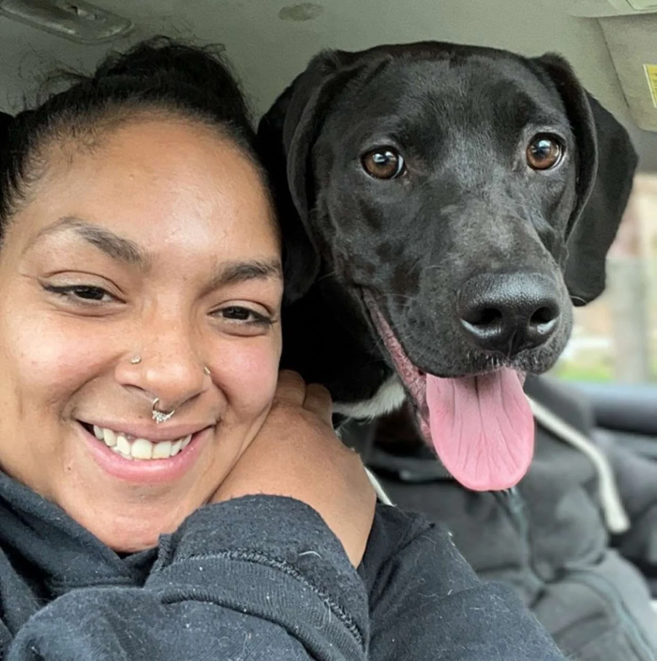 A woman with a nose piercing smiling and a black dog with its tongue out inside a car