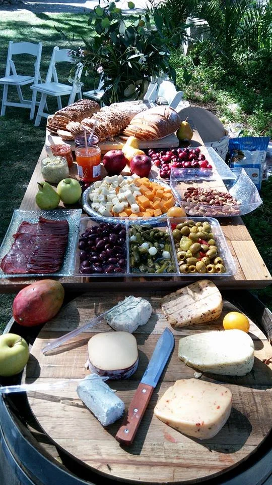 Outdoor table set with assorted cheeses, olives, grapes, bread, and condiments, surrounded by white chairs and greenery.