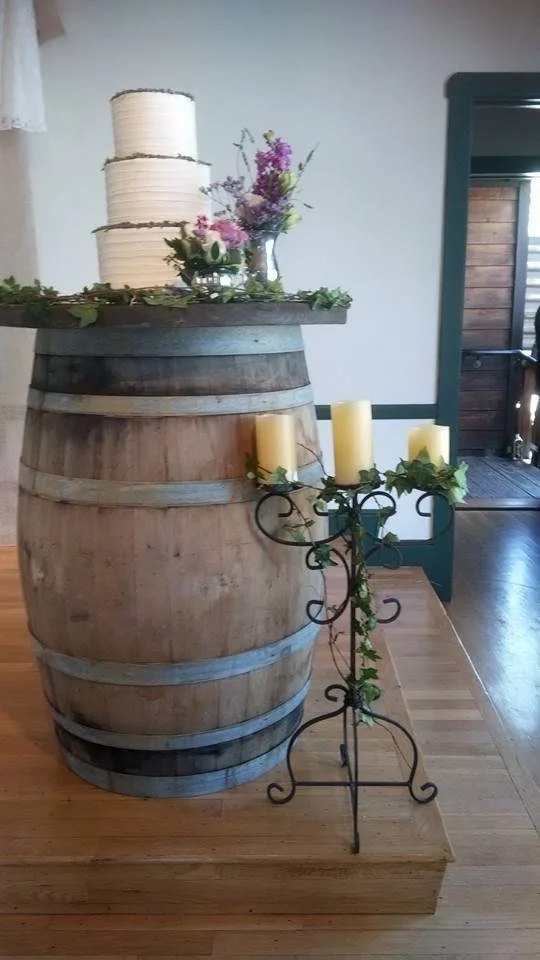 A decorative display with a large wooden barrel, topped with white candles, flowers, and greenery, with a metal candelabra holding four yellow candles with greenery.
