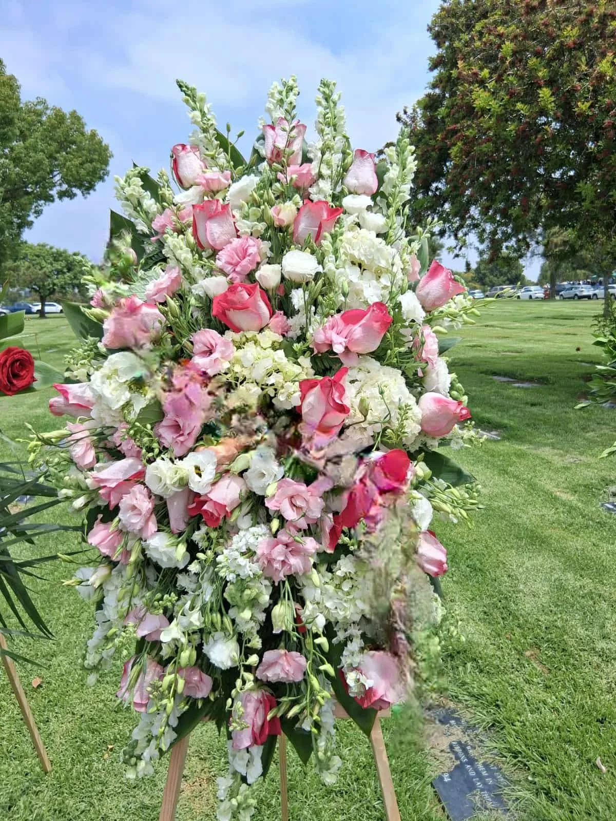 Large floral arrangement with white and pink flowers, including roses and lisianthus, set outdoors in a grassy area with trees and cars in the background.