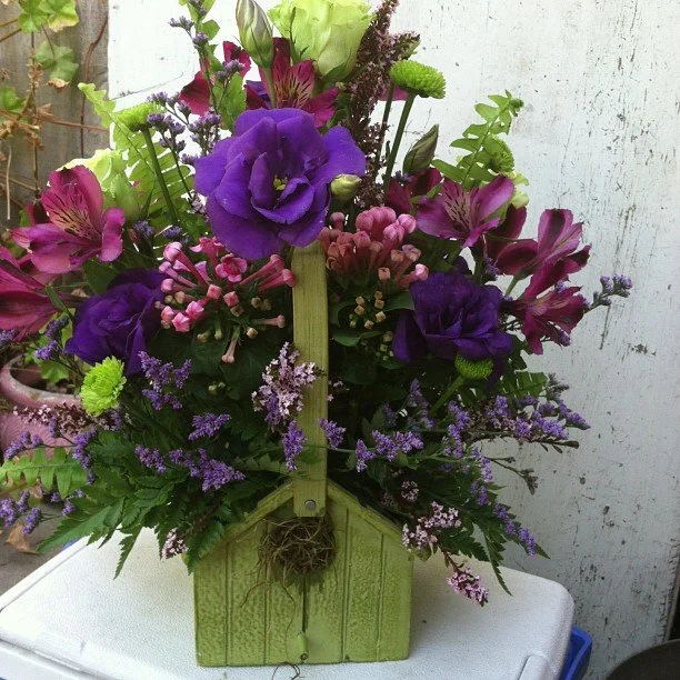 A vibrant floral arrangement in a green wooden container featuring purple lisianthus, pink alstroemeria, lavender, and green accents.