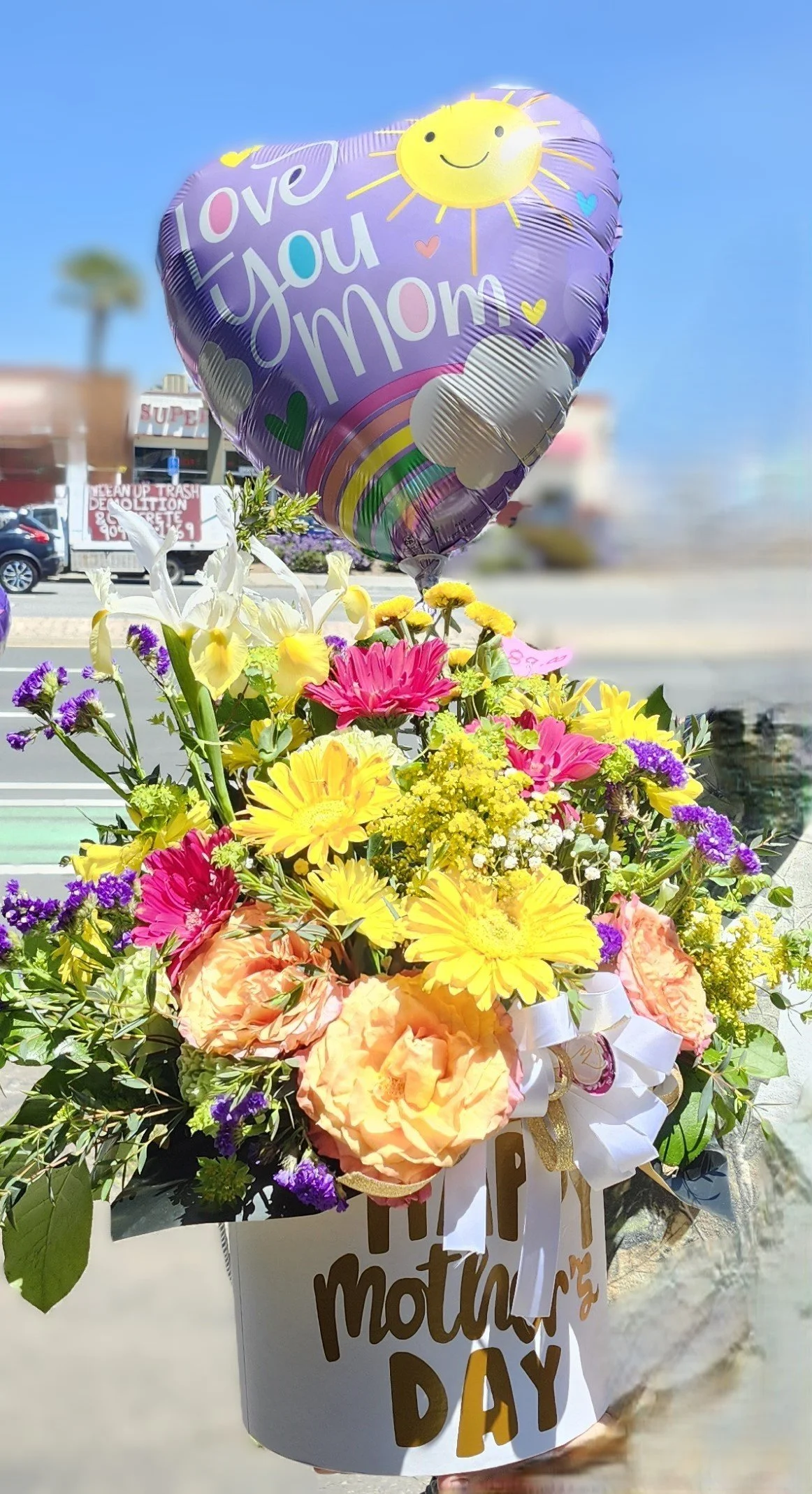 Colorful flower arrangement with yellow, pink, purple, and white flowers in a white box with gold letters reading 'Happy Mother's Day.' A purple heart-shaped balloon above the flowers has a smiling sun and the message 'Love You Mom.'