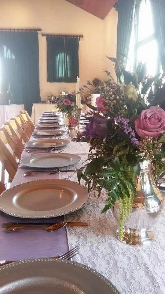 A long dining table set for a meal with plates, silverware, and purple napkins, decorated with flower arrangements in silver vases, in a room with natural light and blue curtains.