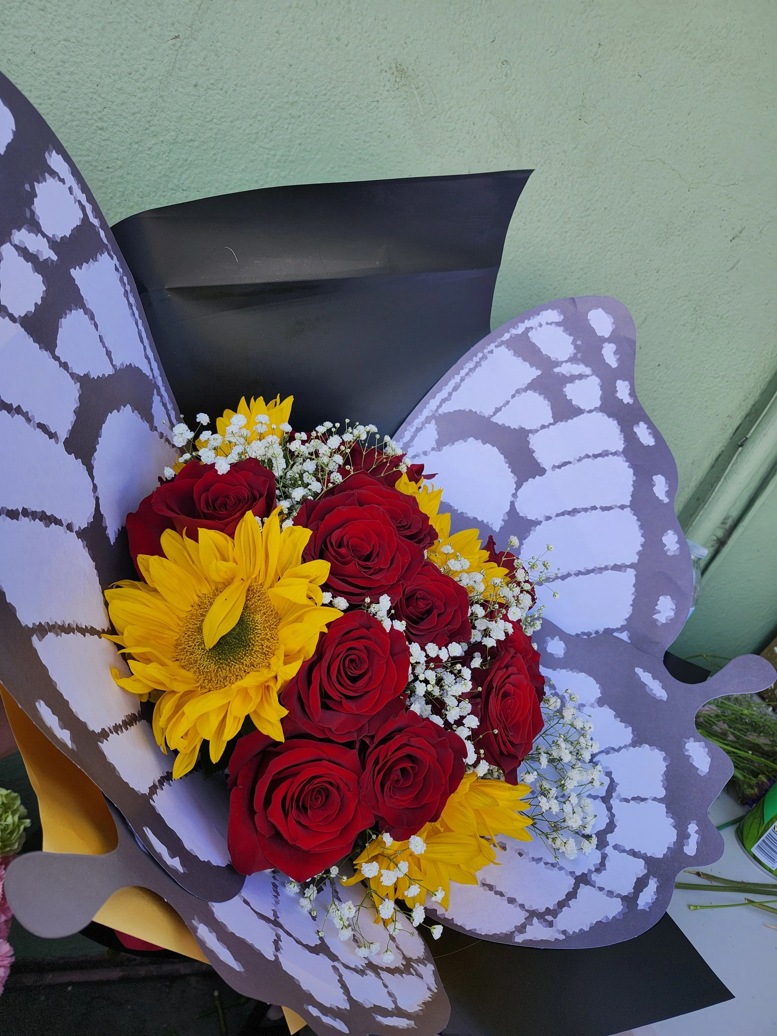 Bouquet of red roses, yellow sunflowers, and small white flowers wrapped in decorative paper.