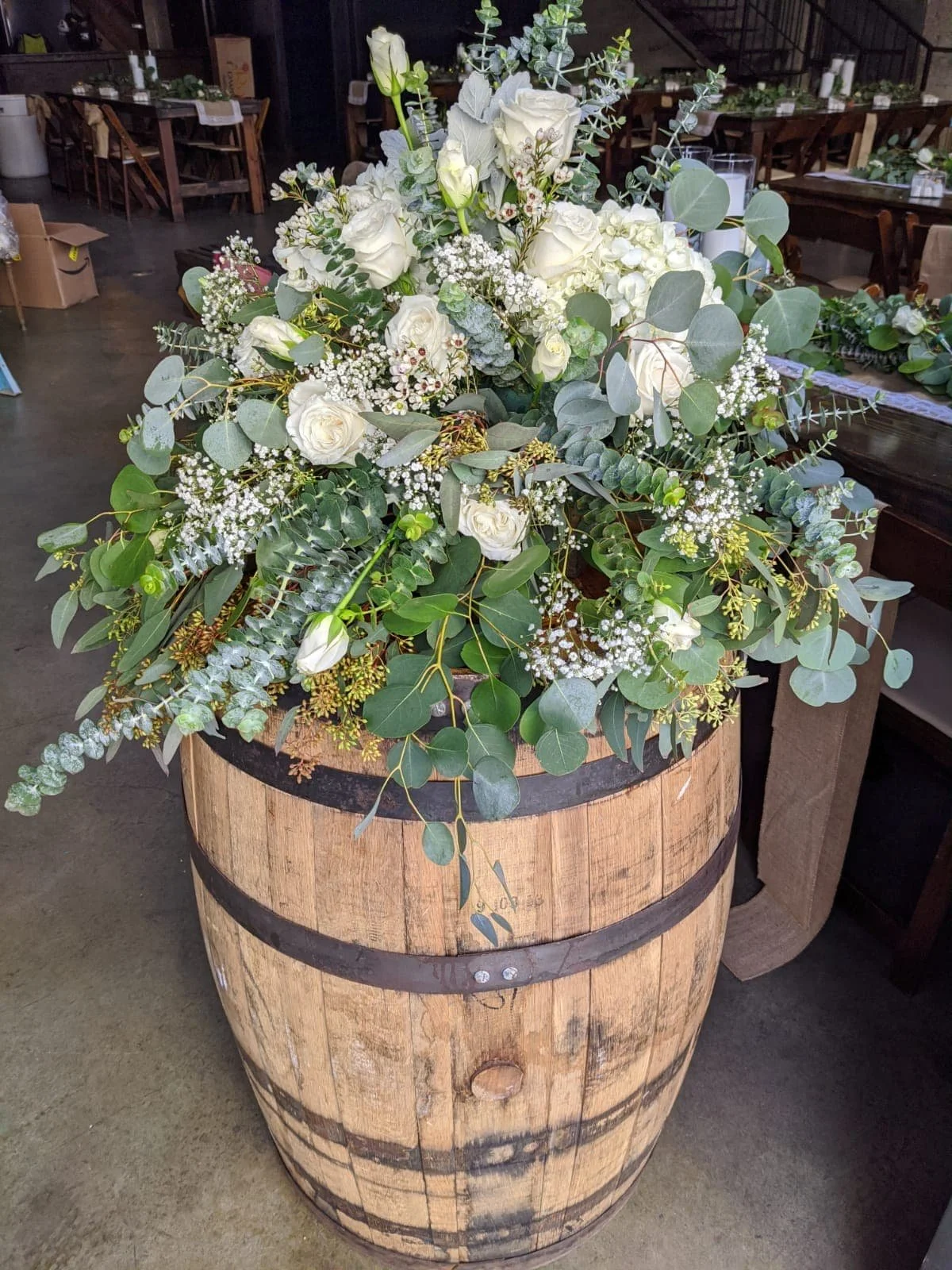 A large floral arrangement with white roses, baby's breath, eucalyptus, and other greenery in a wooden barrel planter.