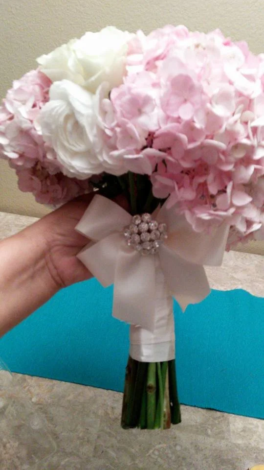 Hand holding a bouquet of pink and white hydrangeas decorated with a white ribbon and pearl embellishment.
