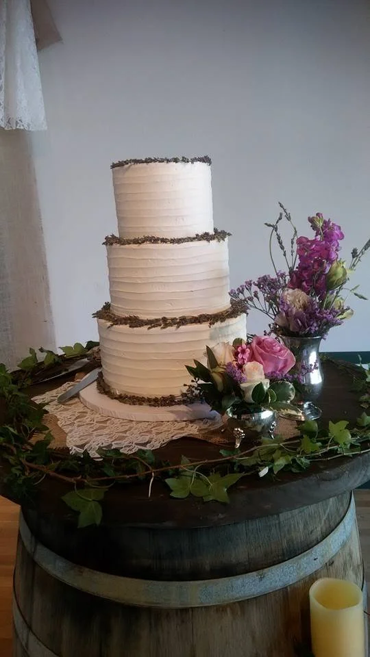 Three-tiered white wedding cake with purple floral decorations on top, placed on a wooden barrel with floral arrangements and candles surrounding it.