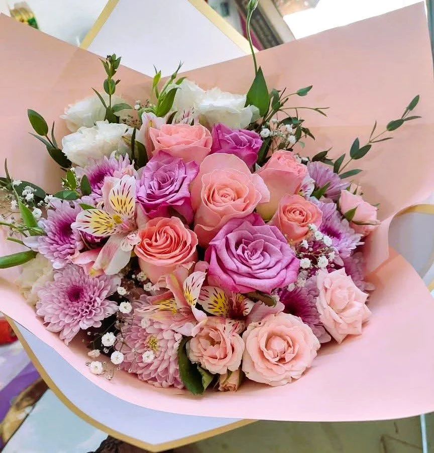 A bouquet of pink, purple, and white roses, adorned with sprigs of baby's breath and green leaves, wrapped in pink and white paper with gold trim.