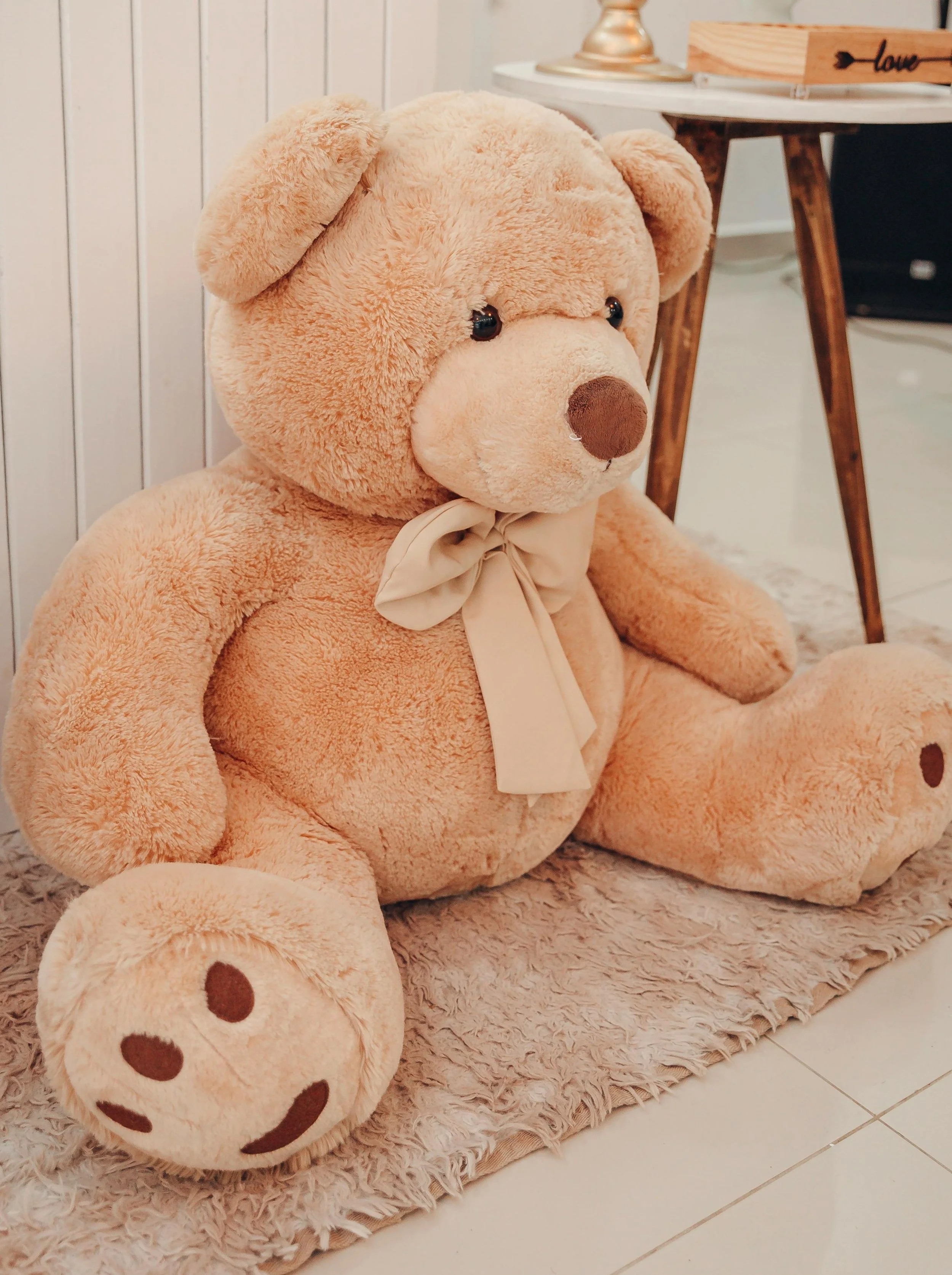 A large beige teddy bear with a cream bow sitting on a fluffy pink rug, with a wooden table and a lamp in the background.