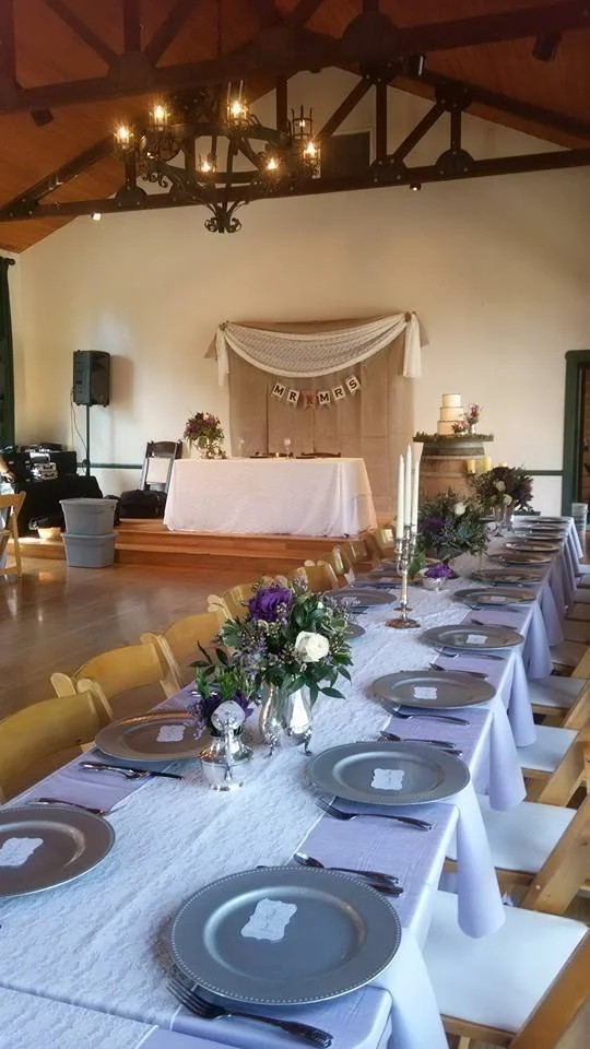 Wedding reception table with plates, cutlery, napkins, and floral centerpieces, with a decorated backdrop and chandelier in a rustic venue